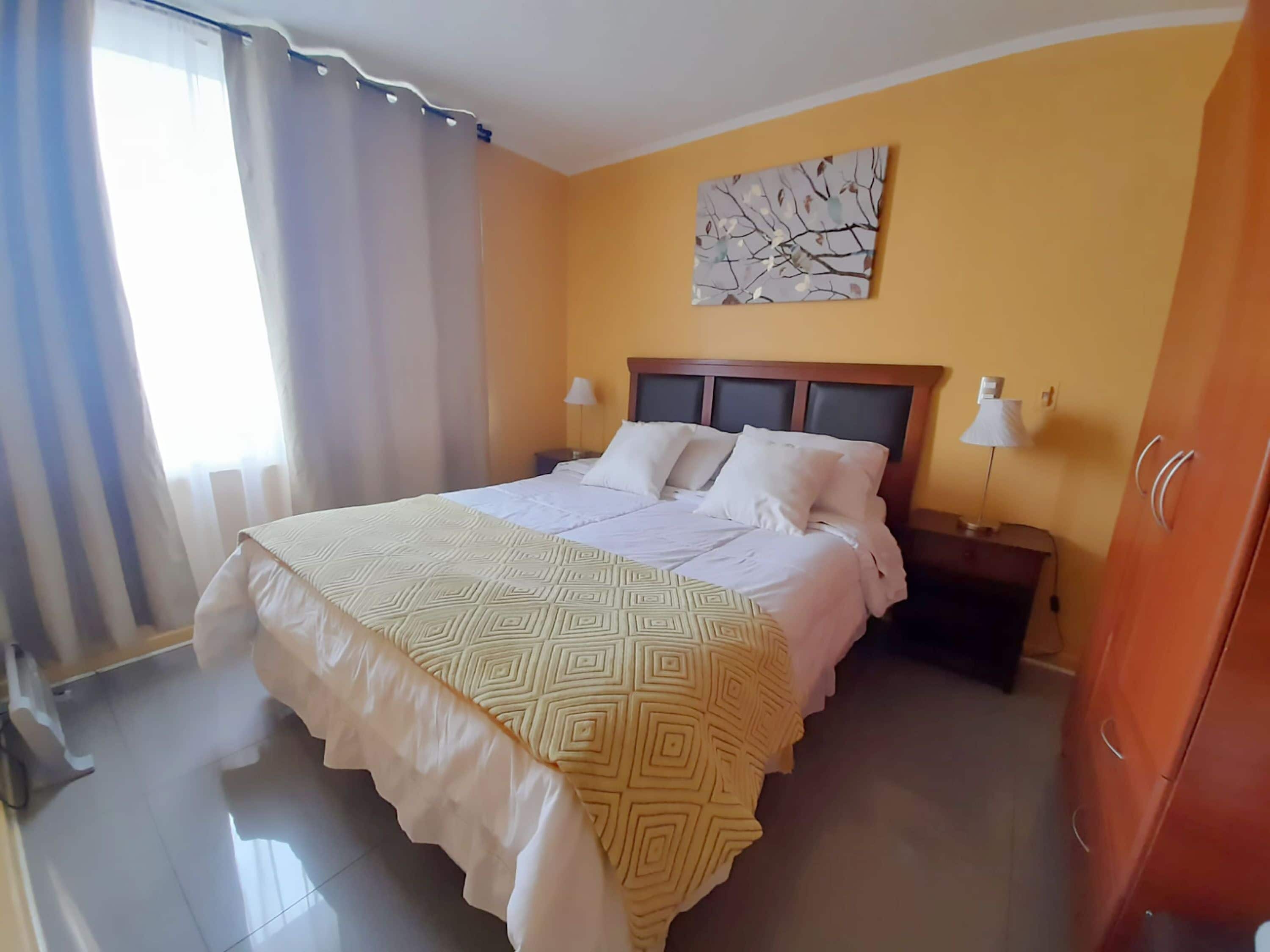 A hotel room with a large bed, a nightstand, a lamp, a wardrobe, and a window with curtains.