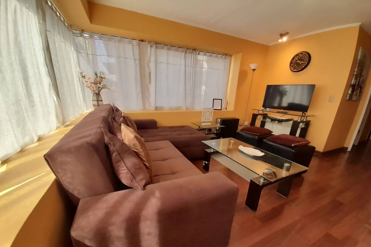 A living room with a brown sofa, a glass coffee table, and a TV on the wall.