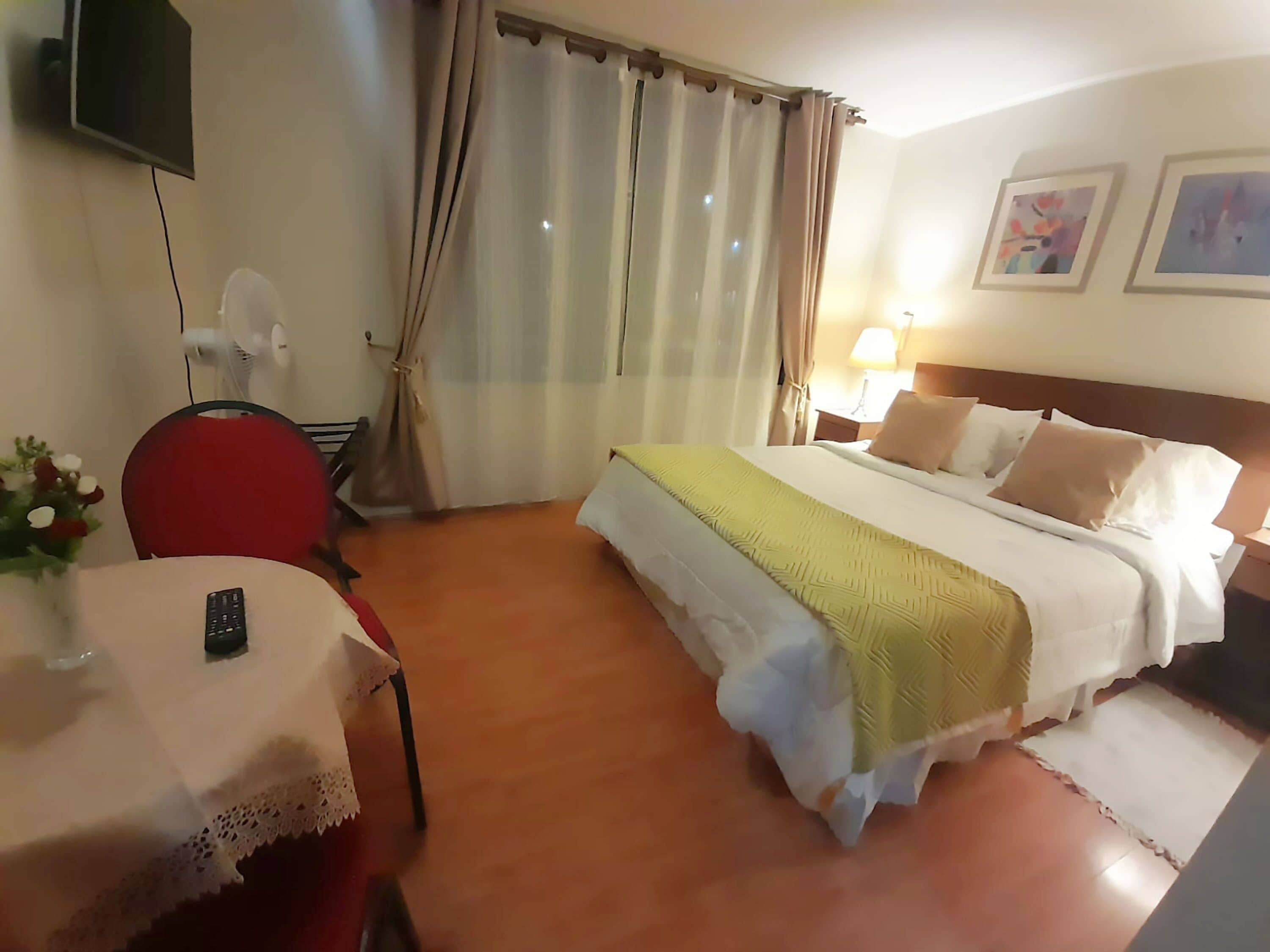 A hotel room with a bed, a desk with a chair, a TV mounted on the wall, and a window with curtains.