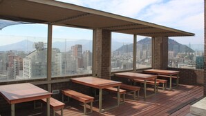 Terraza en la azotea con mesas y bancos de madera, y amplios ventanales con vistas a la ciudad.