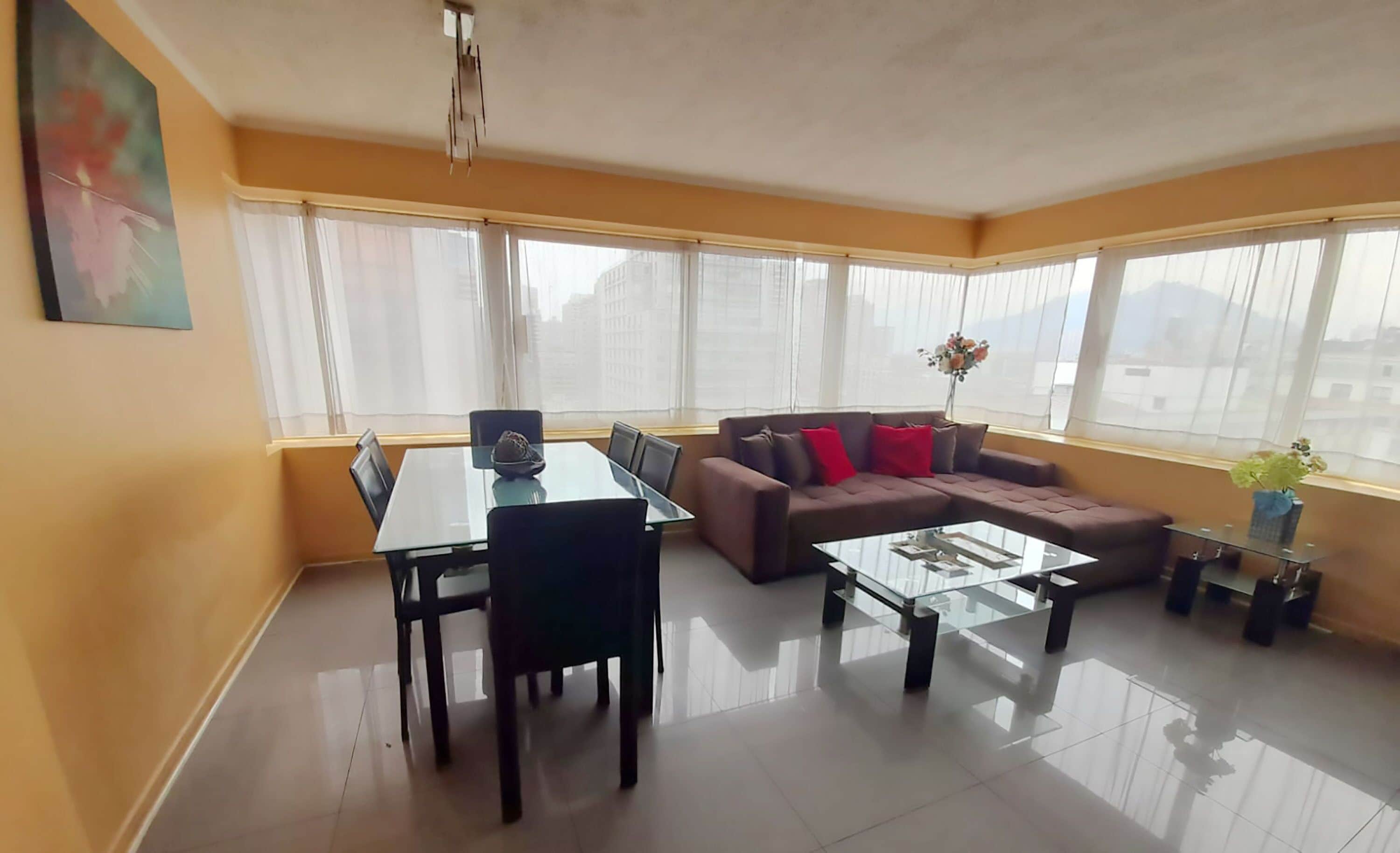A modern living room with a glass coffee table, a brown sofa with red cushions, a dining table with chairs, and large windows with sheer curtains.