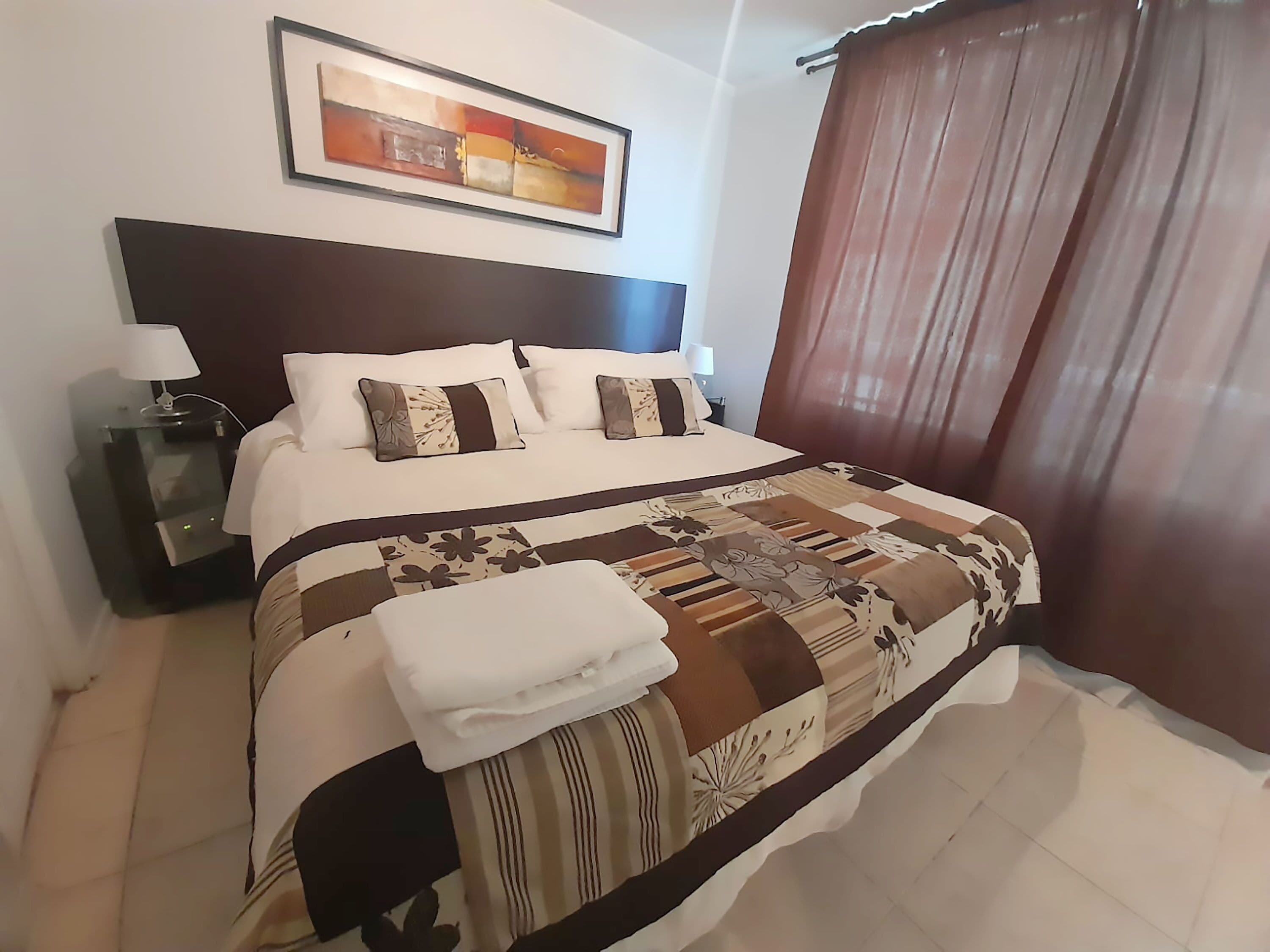 A neatly made bed with a patterned bedspread, a bedside table with a lamp, and a painting on the wall.