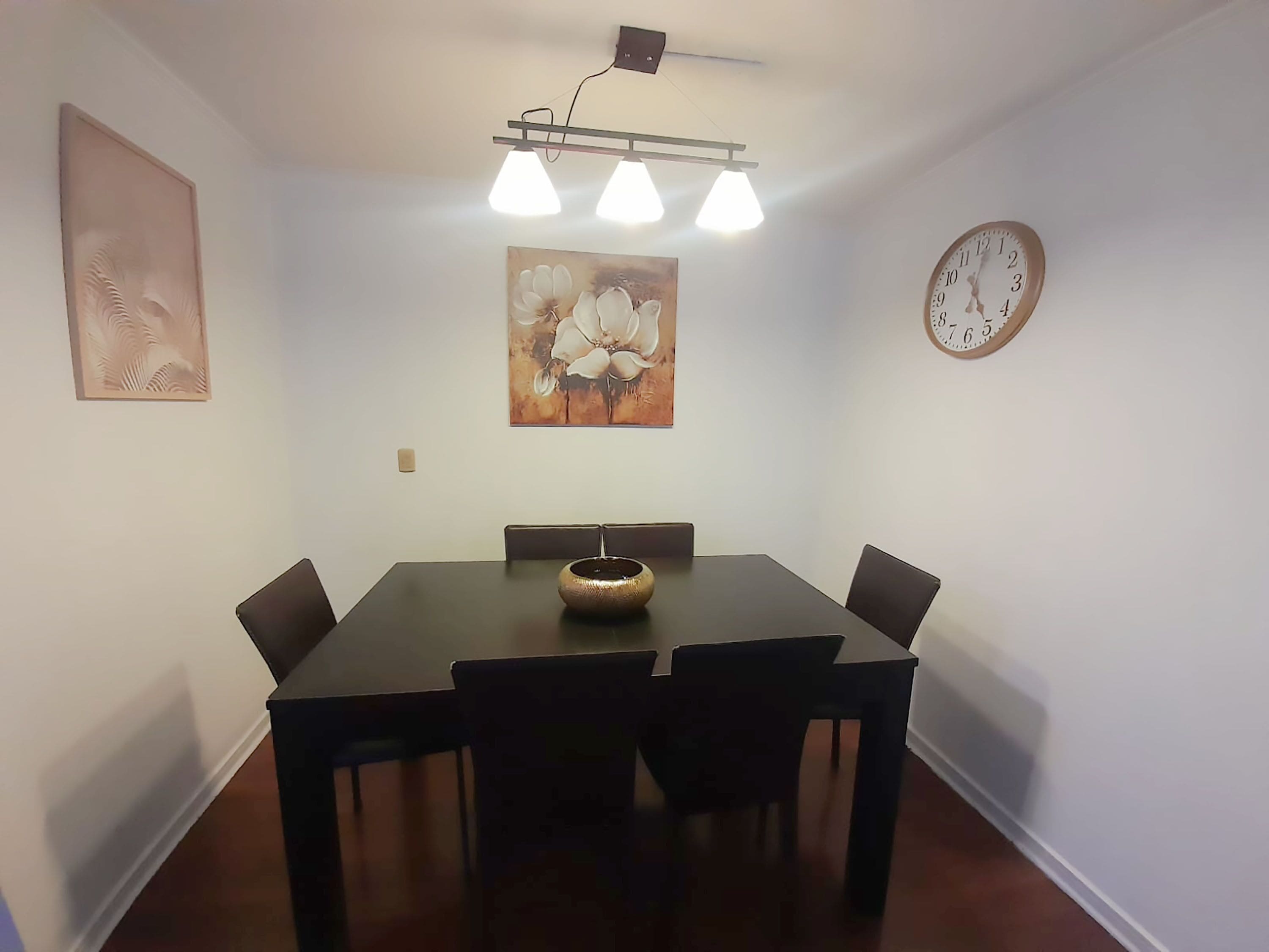 Un comedor con una mesa de madera oscura, cuatro sillas, un jarrón central, un cuadro, un reloj y una lámpara en el techo.