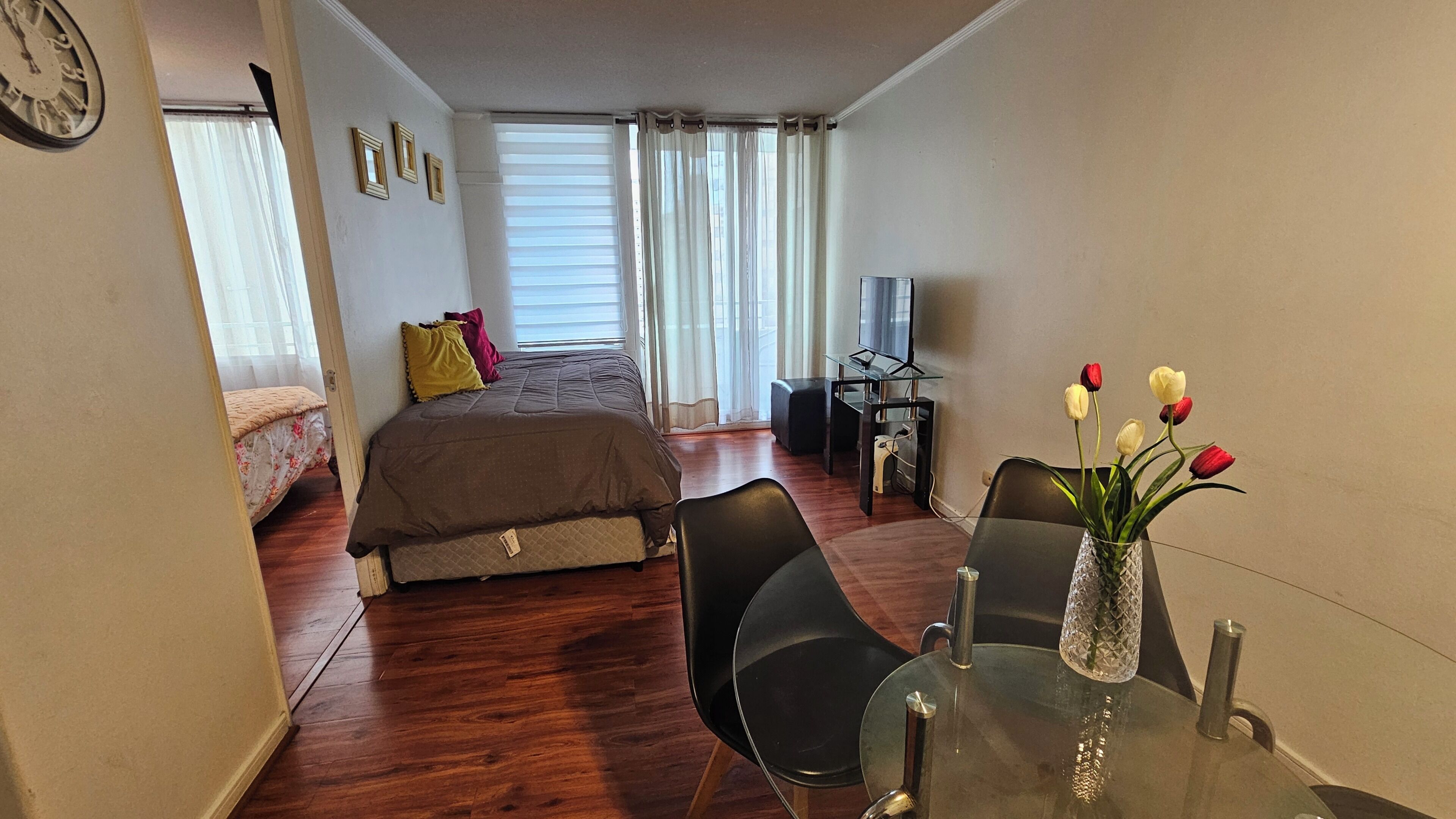 A bedroom with a bed, a desk, a chair, a TV, and a vase with flowers.