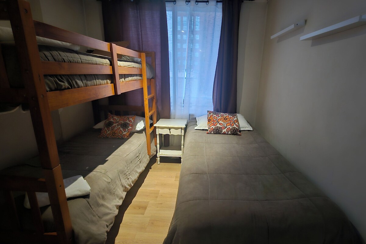 Habitación tipo dormitorio con literas, una mesita de noche pequeña y una ventana con cortinas.