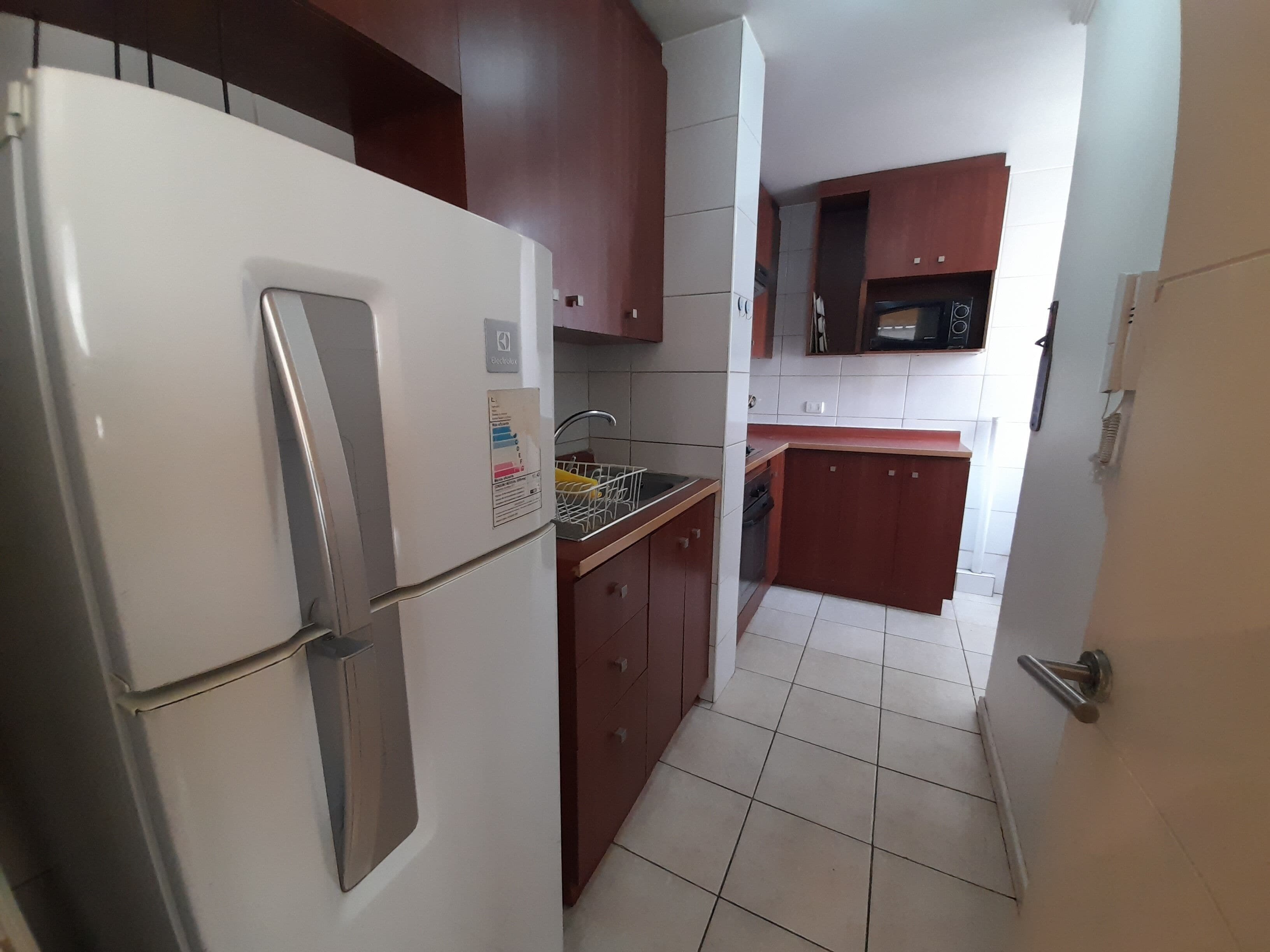 Cozinha compacta com geladeira branca, armários de madeira e piso ladrilhado.
