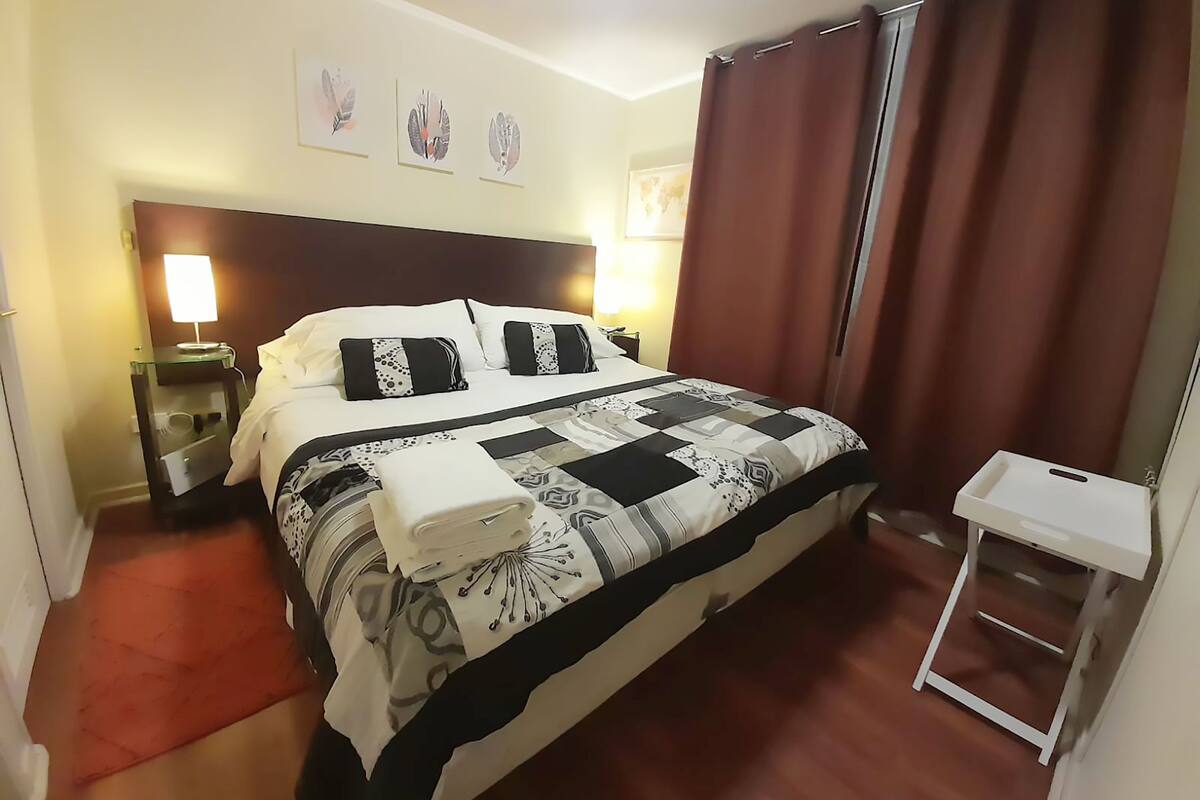 A hotel room with a large bed, a bedside table with a lamp, a small table, and a curtain.