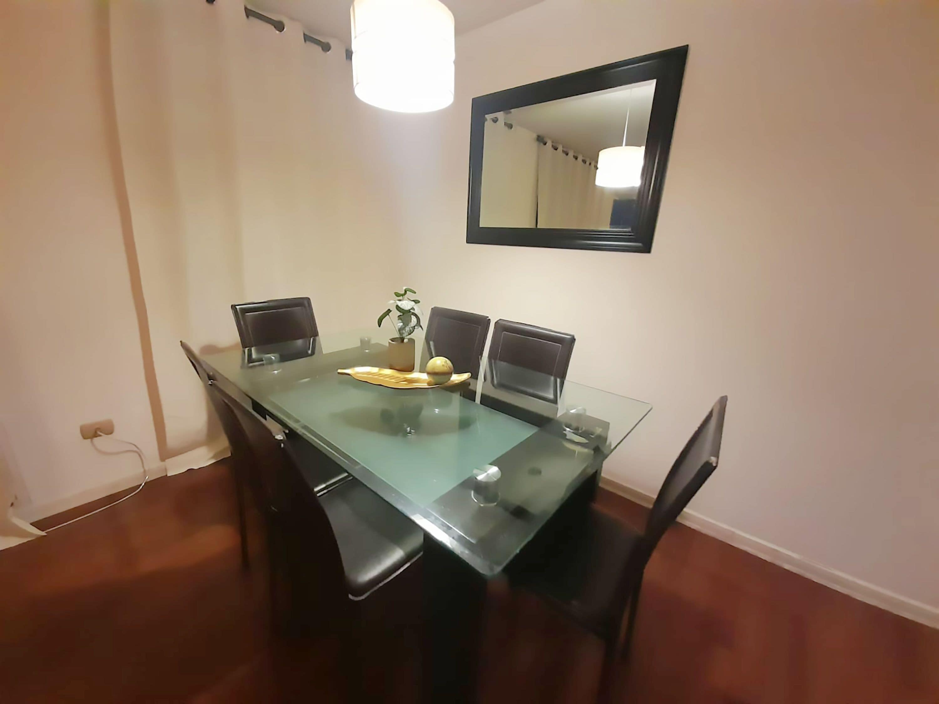 A dining area with a glass table, four chairs, a potted plant, and a mirror on the wall.
