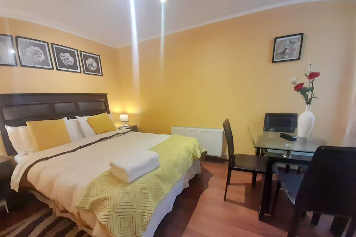 A hotel room with a bed, bedside table, lamp, desk, chair, and a vase with flowers.