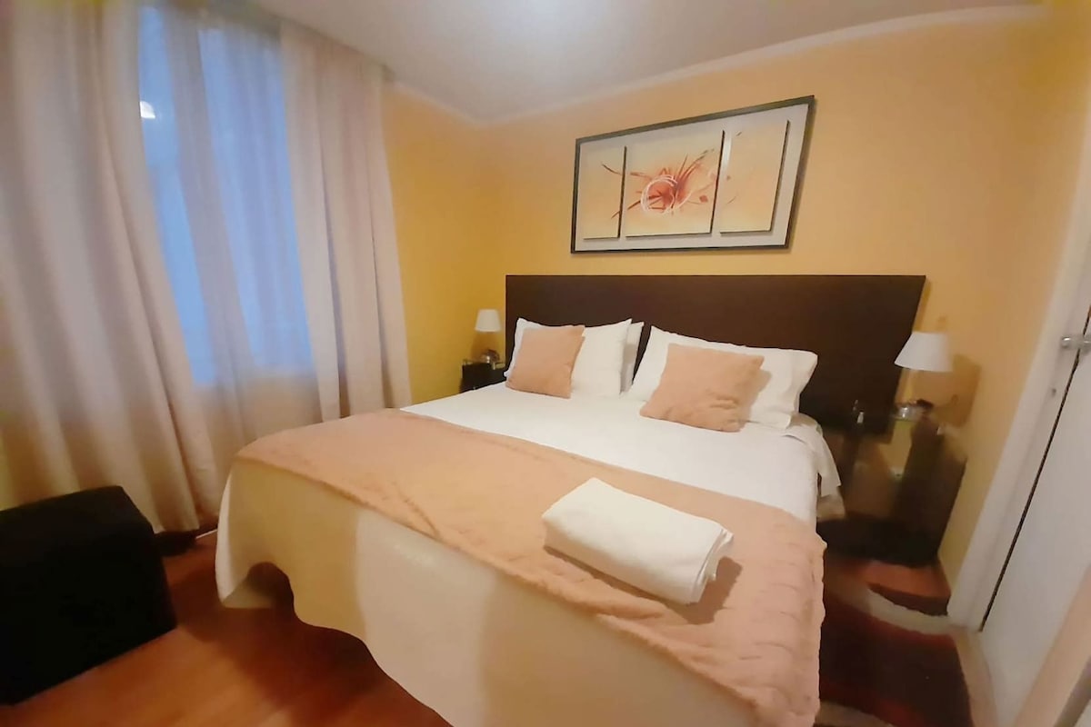 A hotel room with a bed, a nightstand, a lamp, a painting on the wall, and a window with curtains.