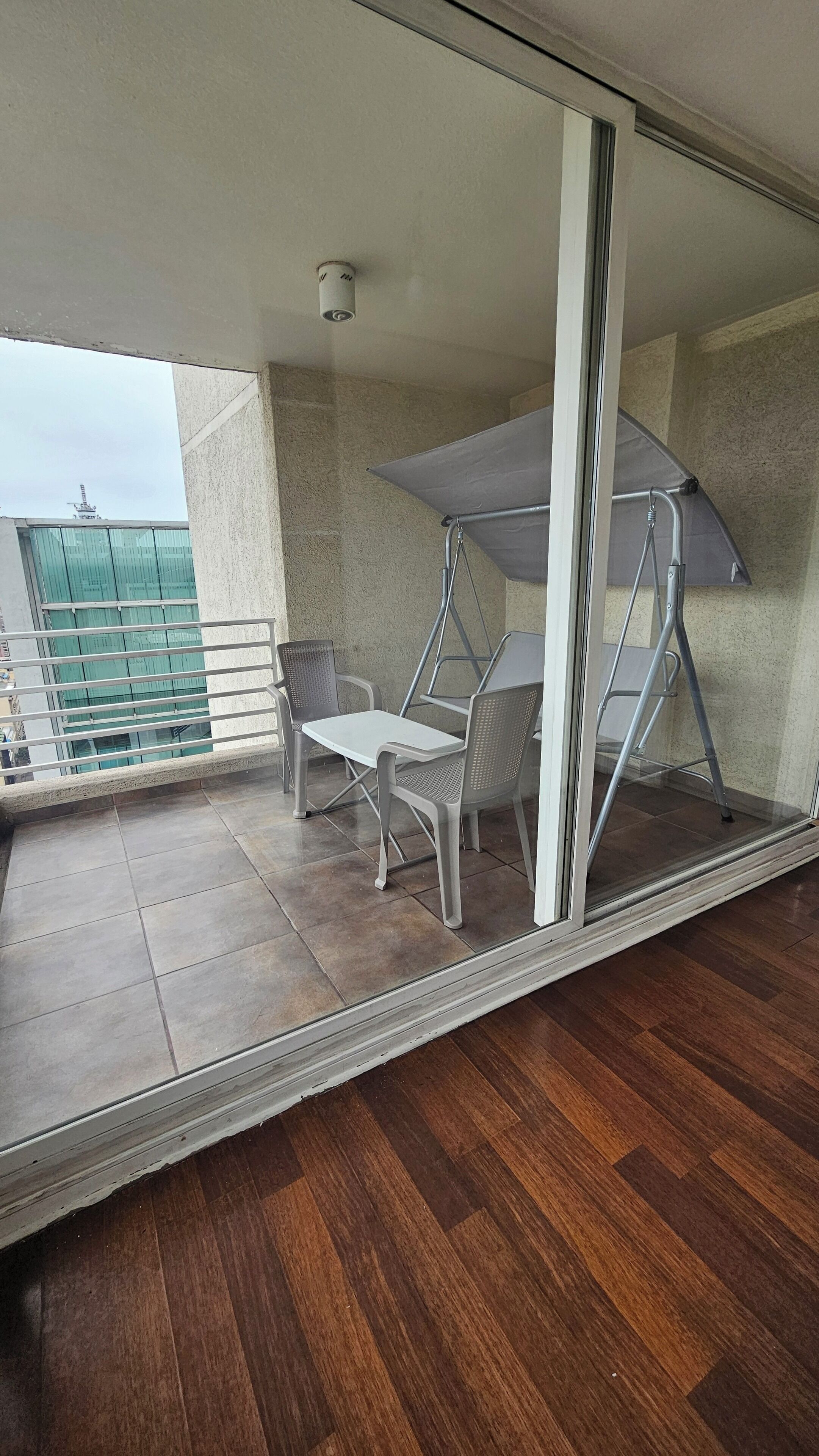 Un balcón con mesa y sillas, un dosel metálico y vista a un edificio de fachada verde.