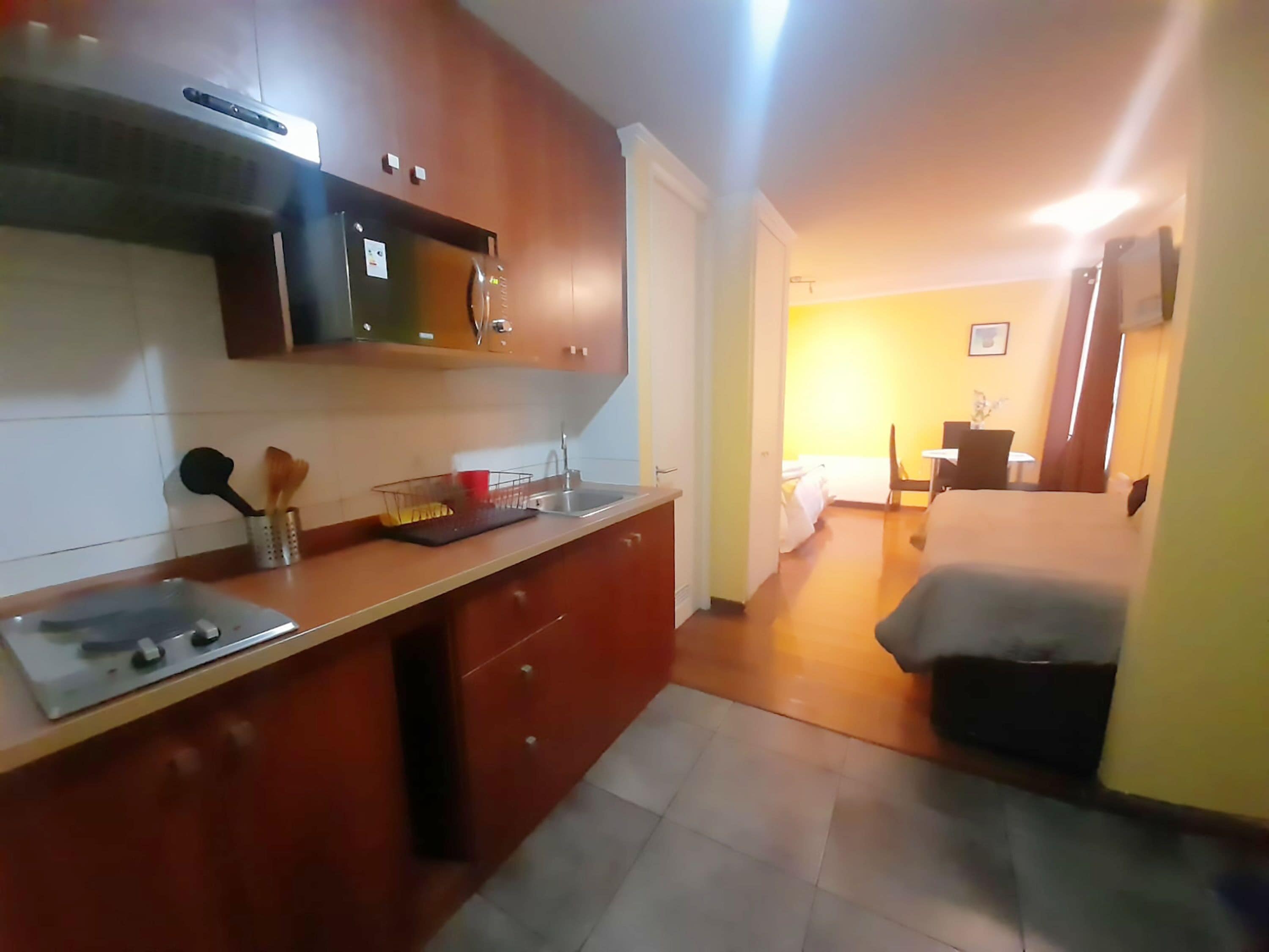 Cocina compacta con microondas, lavabo y estufa. Se ve una zona de comedor con una mesa y sillas al fondo.