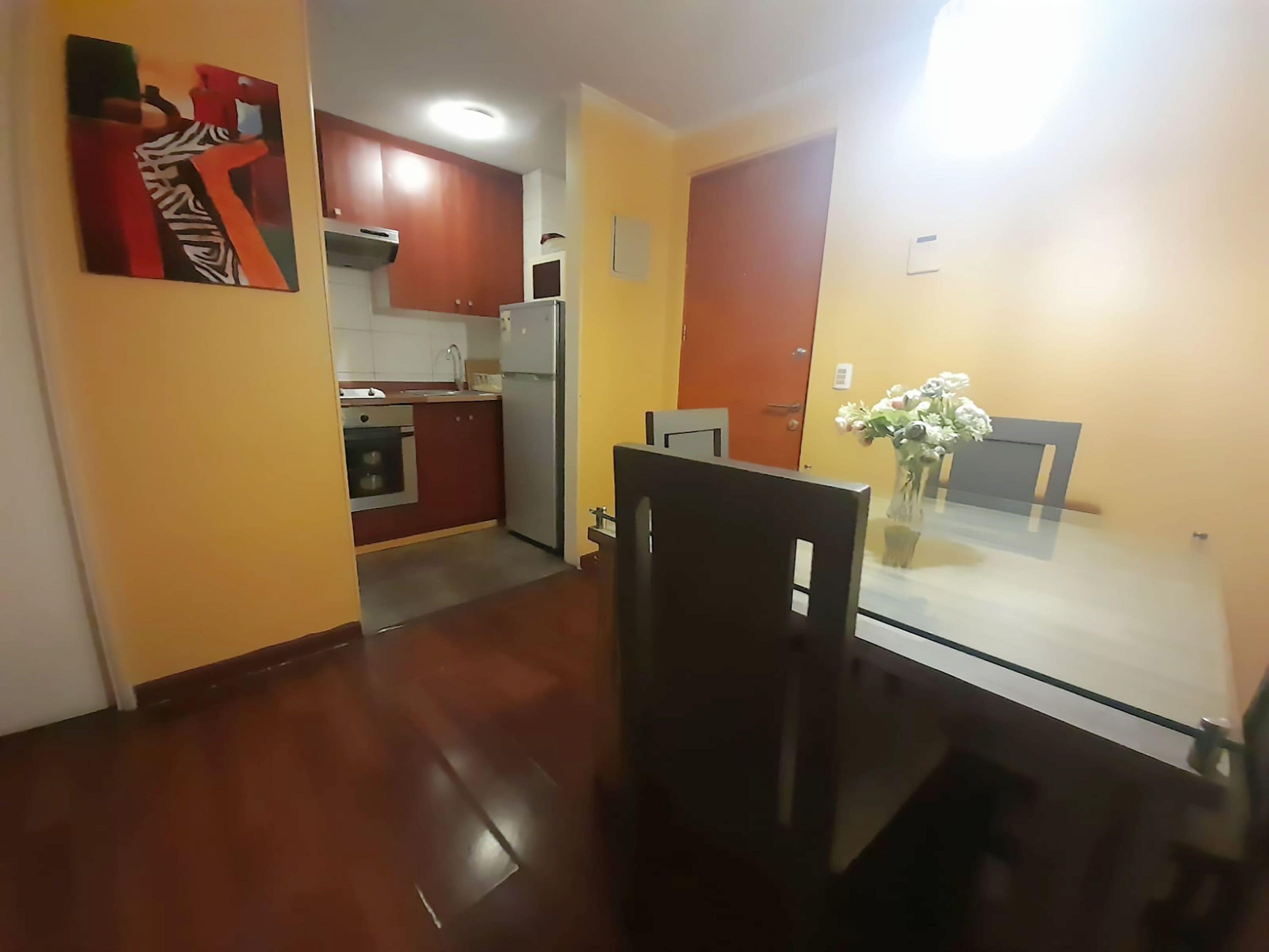 Um espaço de estar compacto com cozinha, área de refeições e uma pequena mesa com um vaso de flores.