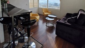 Un salón moderno con un piano, sillas amarillas, un sofá negro y vistas a un paisaje urbano a través de amplios ventanales.