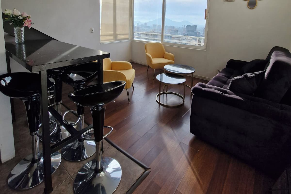 A modern living room with a piano, yellow chairs, a black sofa, and a view of a cityscape through large windows.