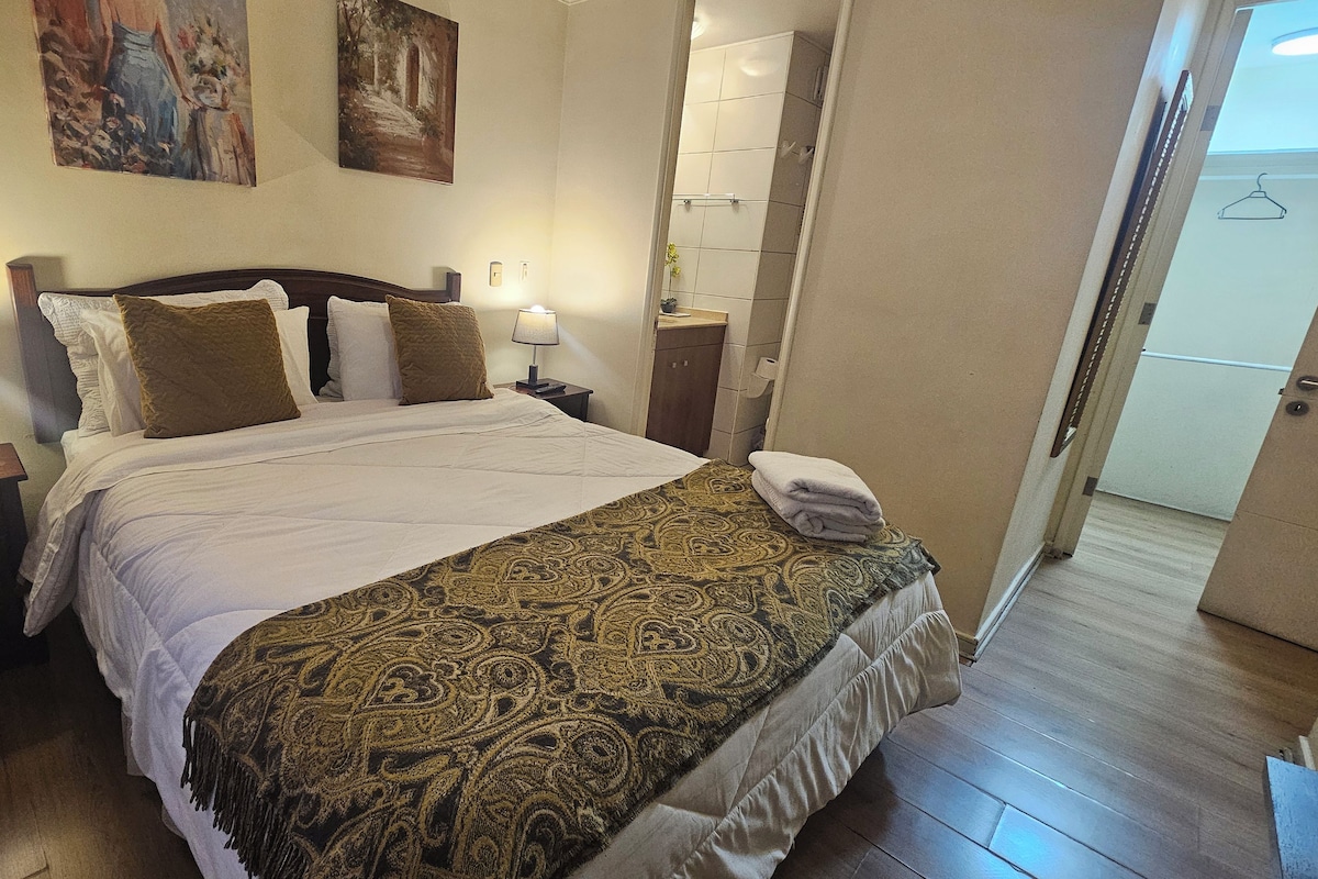 Una cama bien tendida con un cubrecolcha estampado, dos lámparas de noche y una puerta que da a otra habitación.