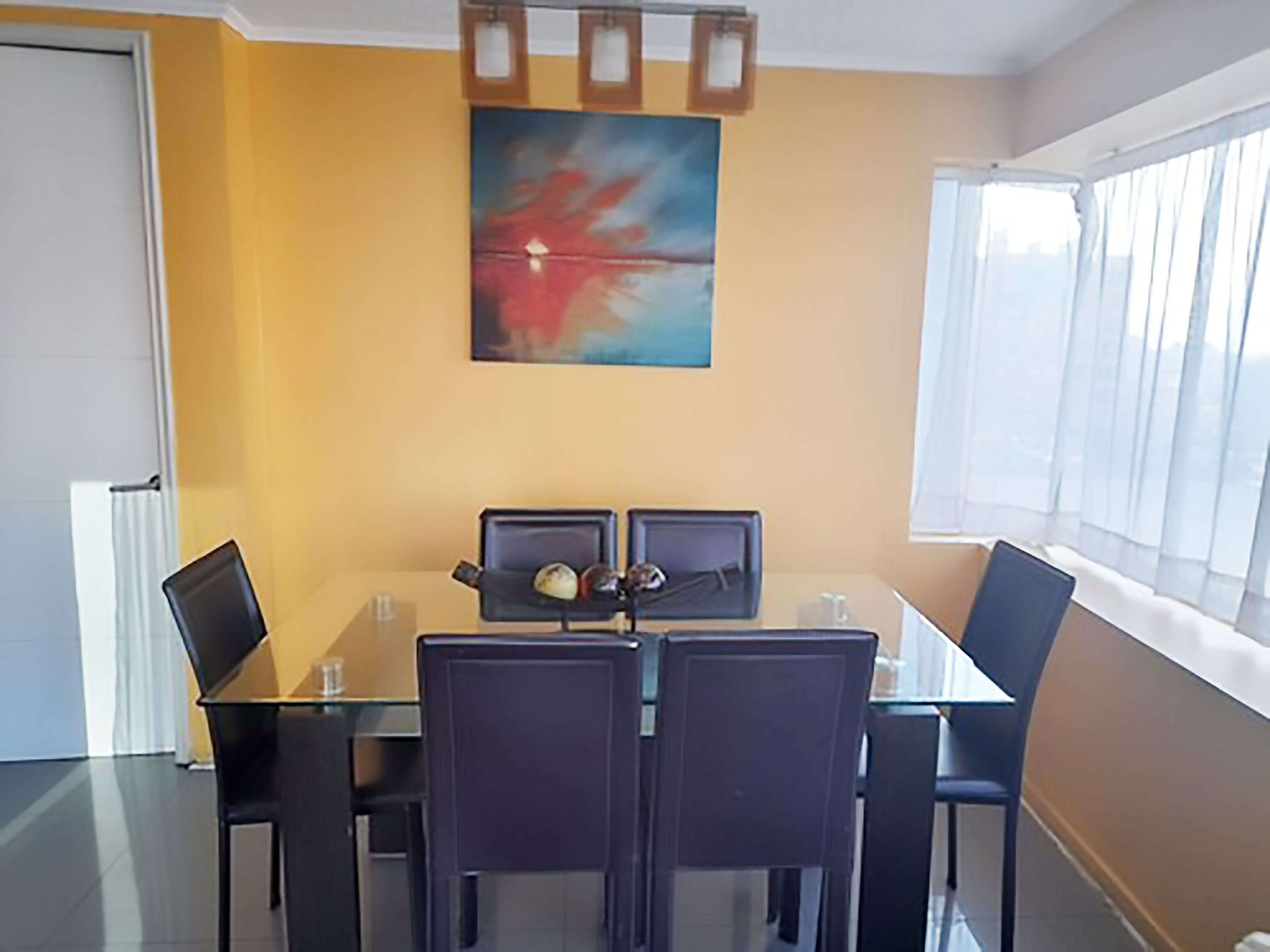 A dining area with a glass table and four chairs, a painting on the wall, and a window with curtains.