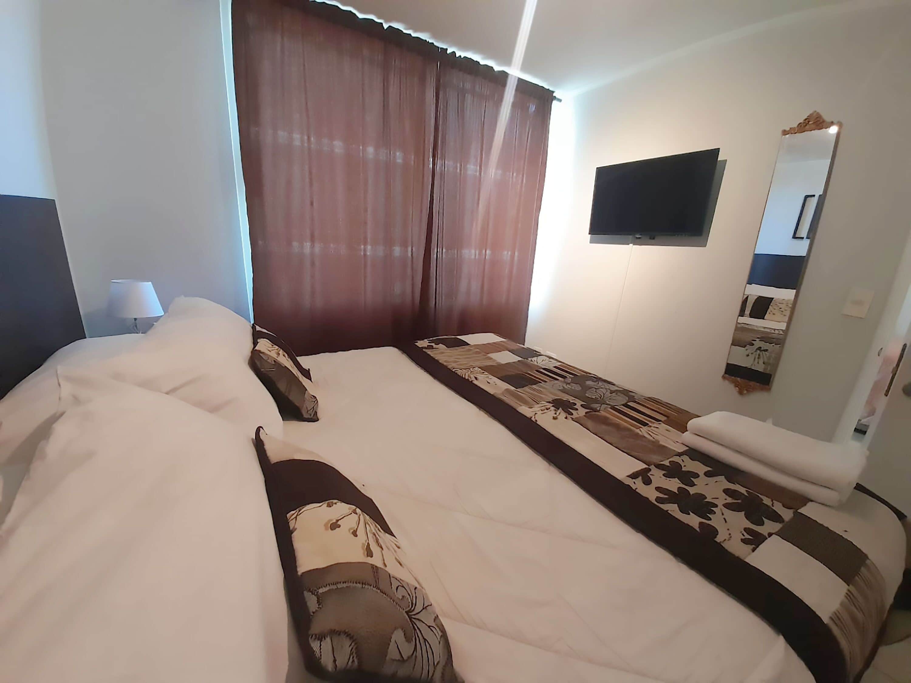 Habitación de hotel con una cama grande, un televisor de pantalla plana montado en la pared, una mesita de noche con lámpara y una ventana con cortinas.