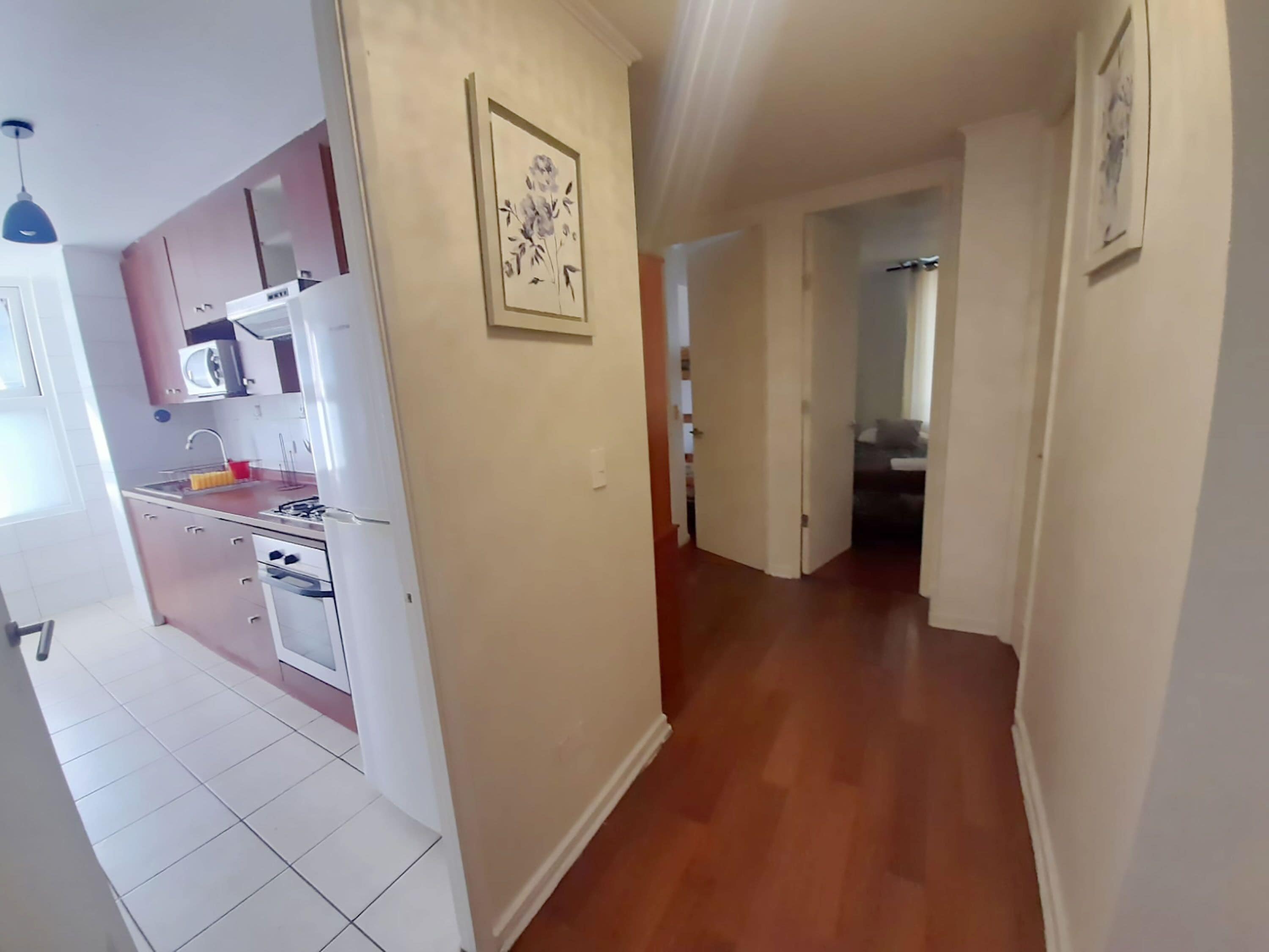 A kitchen with white cabinets, a microwave, and a sink. There is a hallway with wooden flooring leading to a bedroom.