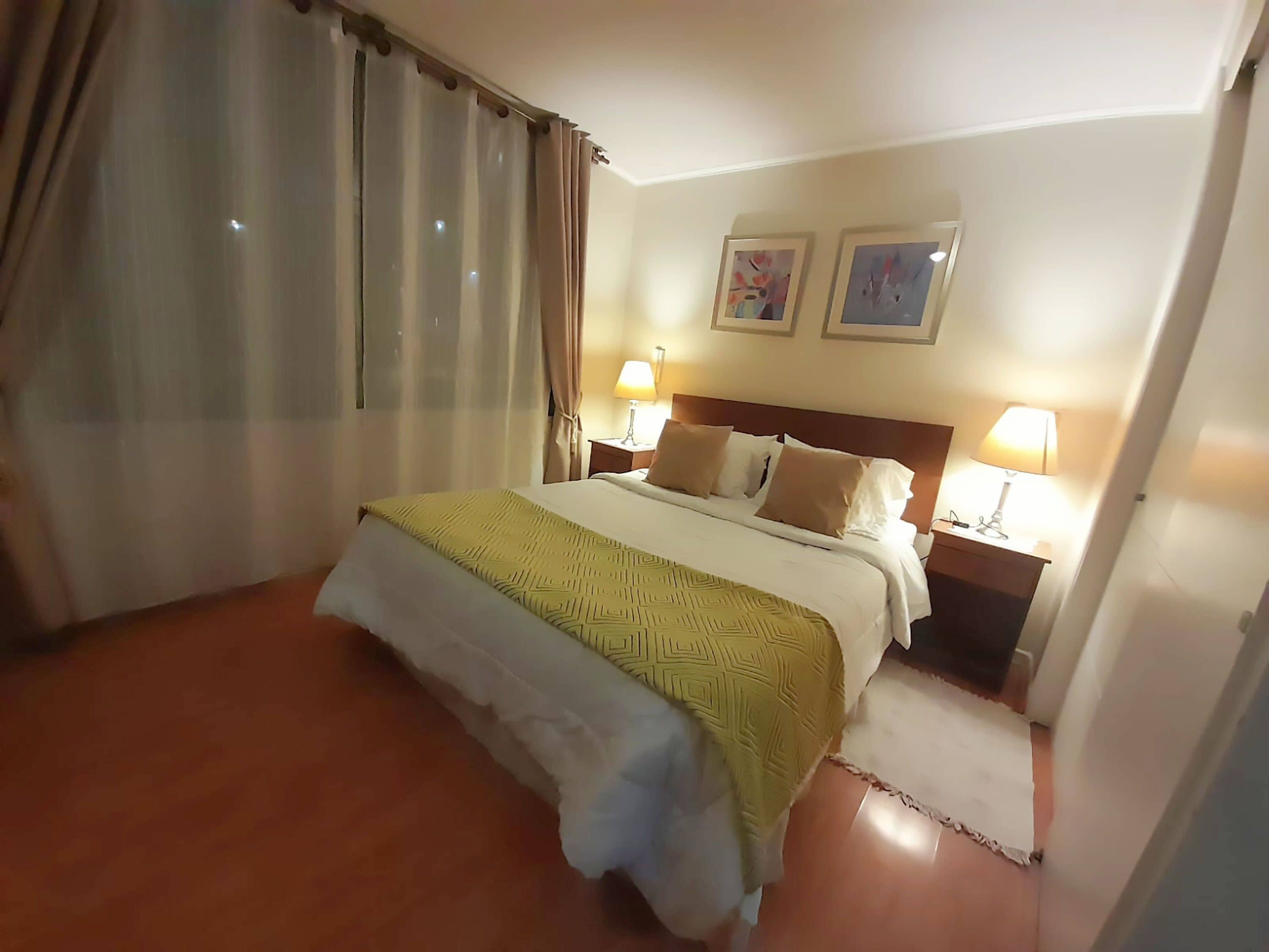 A hotel room with a bed, two bedside lamps, a headboard, and a window with curtains.