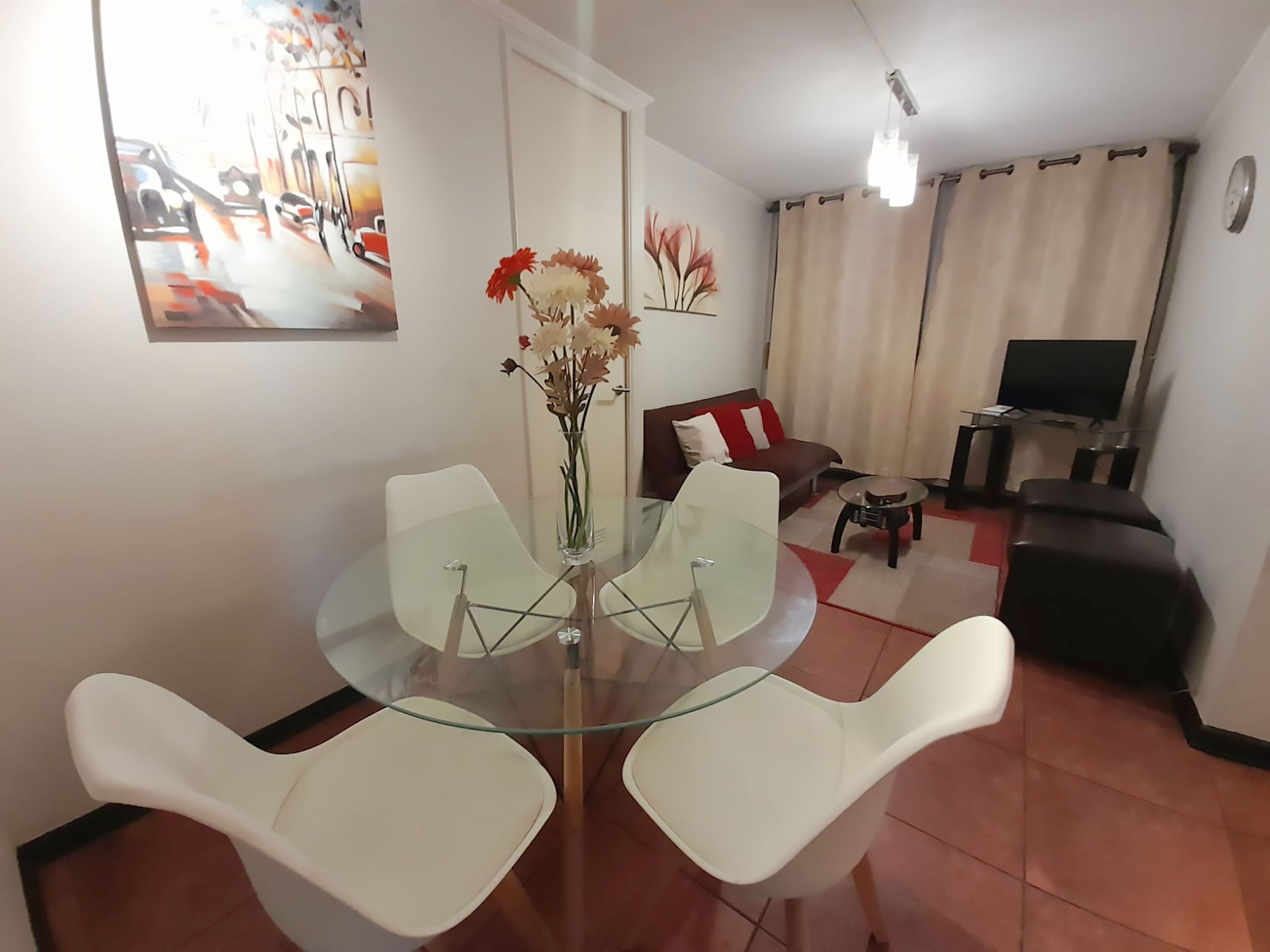 Una sala de estar moderna con una mesa de comedor de vidrio, sillas blancas, un sofá, un televisor y un cuadro en la pared.