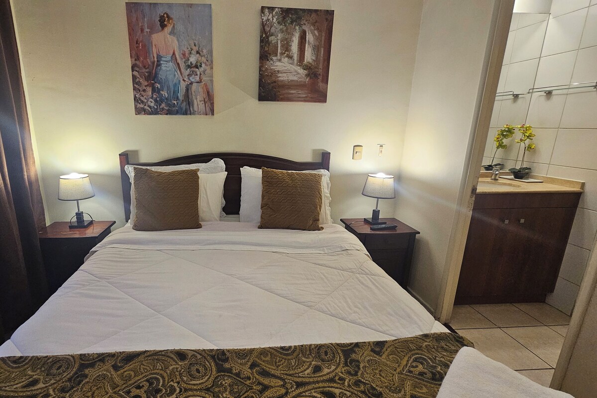 Una habitación de hotel con cama, dos cuadros en la pared, una mesita de noche con lámpara y un baño con lavabo y espejo.