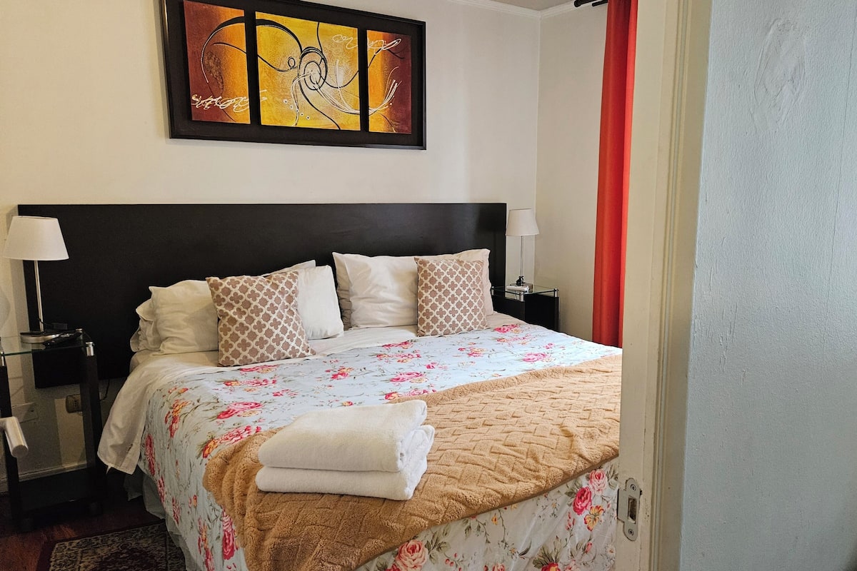 A hotel room with a bed, pillows, a nightstand, a lamp, and a painting on the wall.
