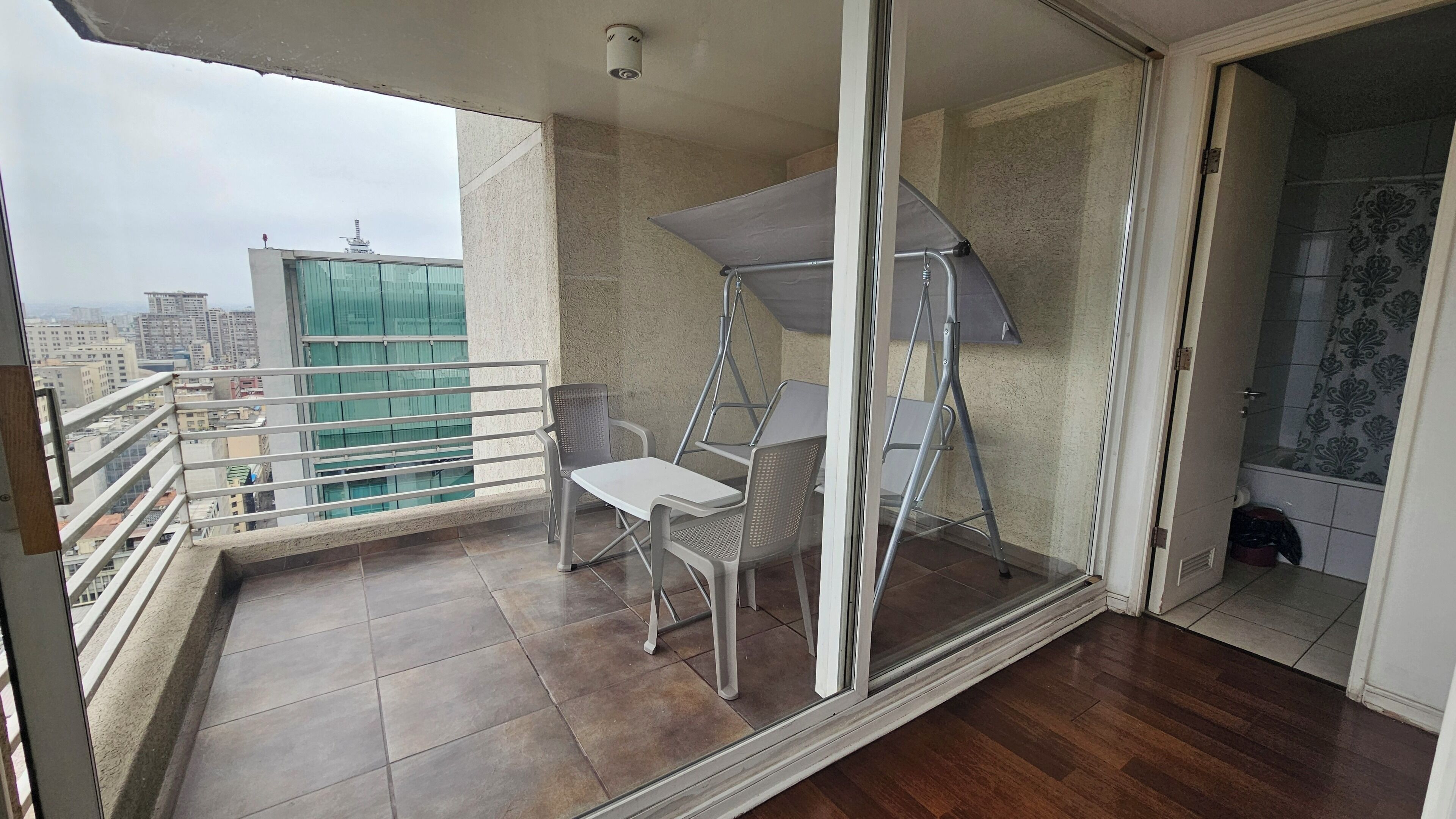 Un balcon avec une table et des chaises blanches, une balançoire en métal et une vue sur les immeubles de la ville.