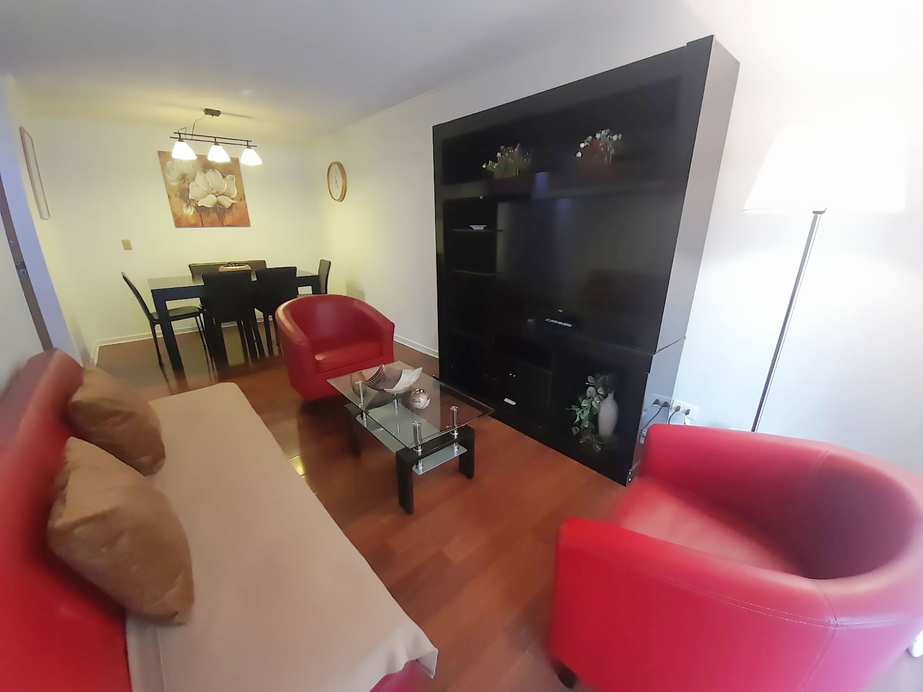 A living room with a sofa, two red armchairs, a coffee table, a black entertainment unit, a dining area with a table and chairs, and a clock on the wall.