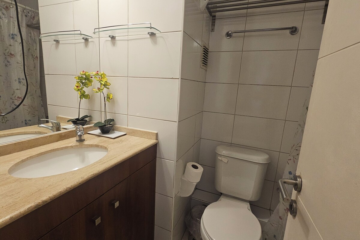 Un baño con lavamanos doble, inodoro, ducha con puerta de vidrio y espejo con cortina estampada.