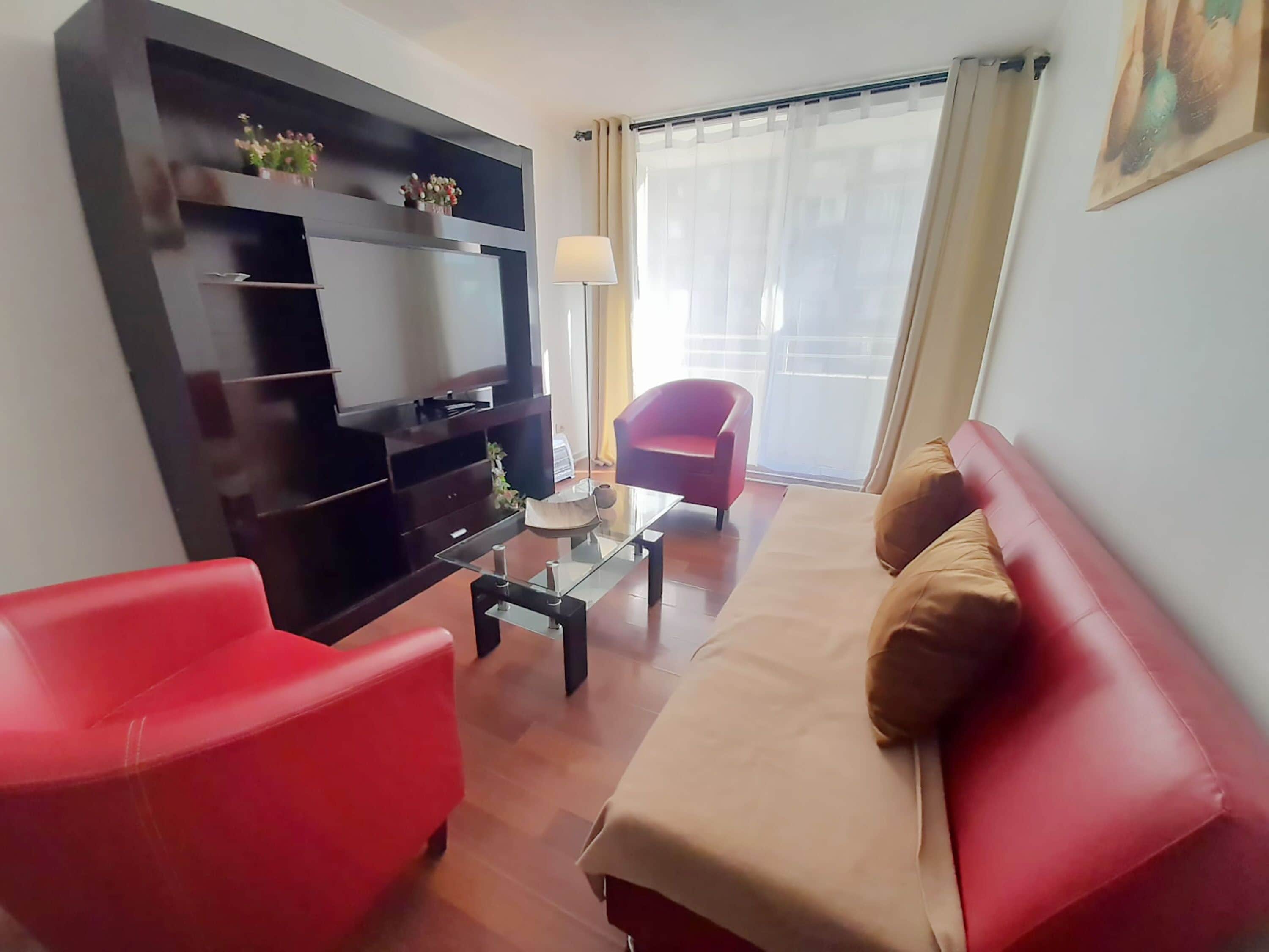 A modern living room with a sofa, two red armchairs, a glass coffee table, a TV on a black stand, and a window with curtains.