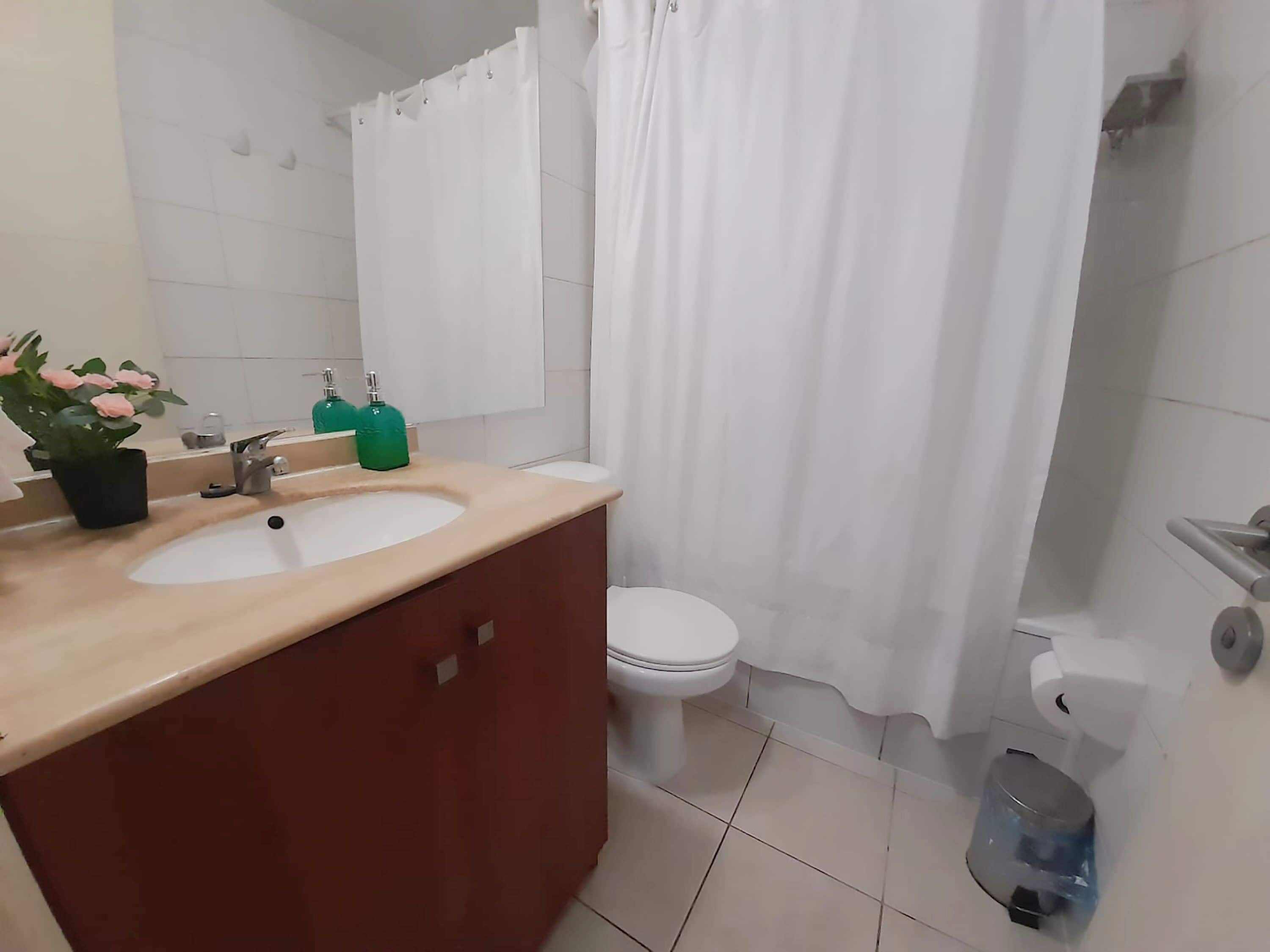 A bathroom with a double sink vanity, a toilet, a shower curtain, and a trash bin.