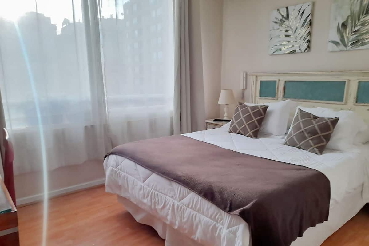 A hotel room with a bed, a nightstand, a lamp, and a view of buildings through sheer curtains.
