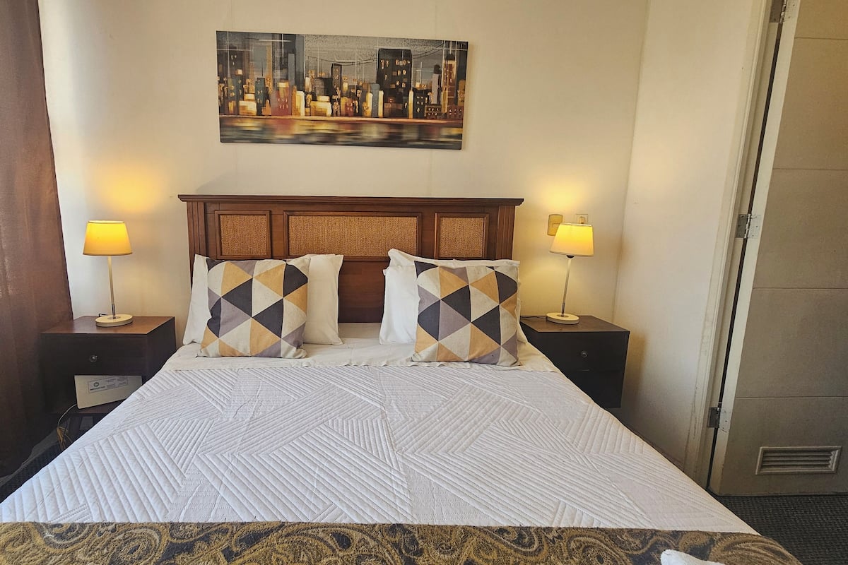 A hotel room with a large bed, two bedside tables with lamps, a headboard, and a cityscape painting on the wall.