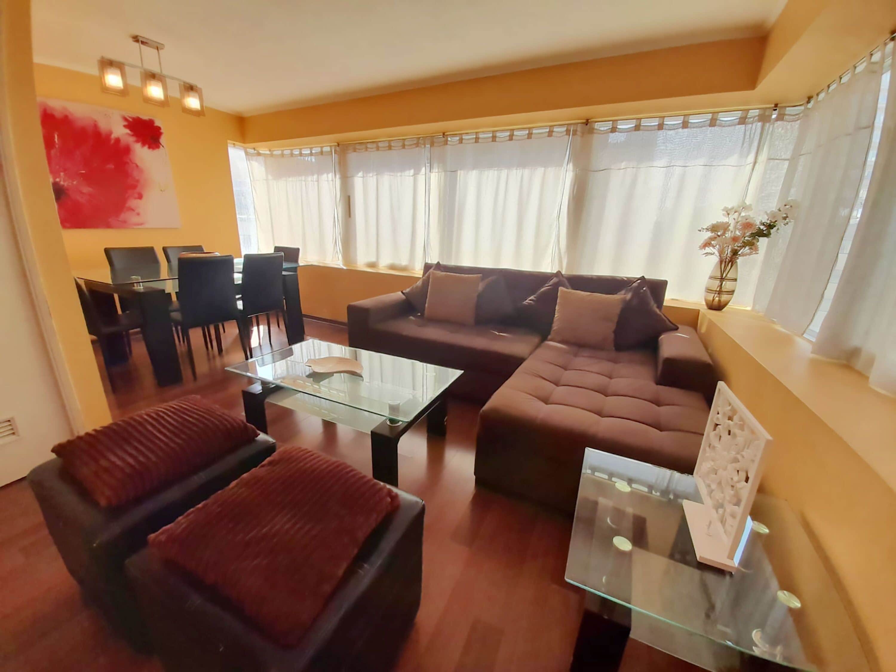 Een moderne woonkamer met een bruine bank, twee fauteuils, een glazen salontafel, en een eethoek met een glazen tafel en stoelen.