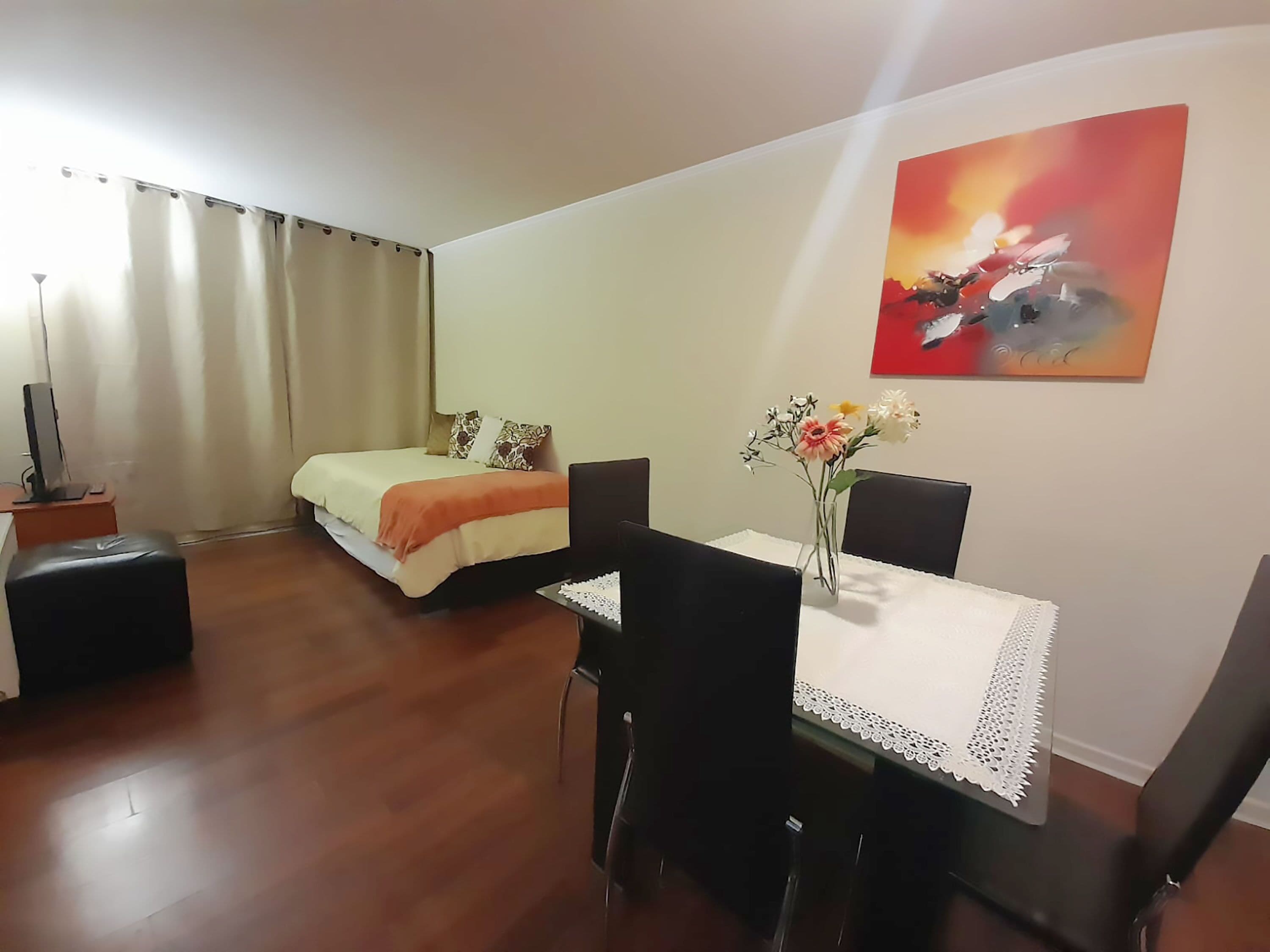 Ein Zimmer mit einem Bett, einem Esstisch mit Stühlen, einer Blumenvase und einem Bild an der Wand.