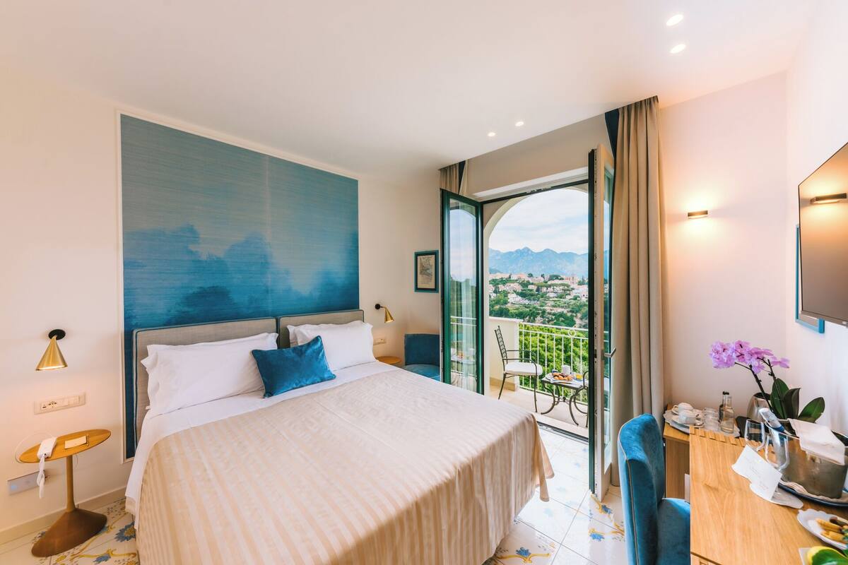 Une chambre d’hôtel avec un lit, un balcon donnant sur un beau paysage, une table ornée d’un vase de fleurs et un tableau accroché au mur.