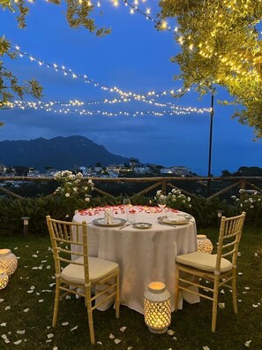 An outdoor dining area with a round table set for four, surrounded by chairs, under string lights, with a view of a mountainous landscape in the background.