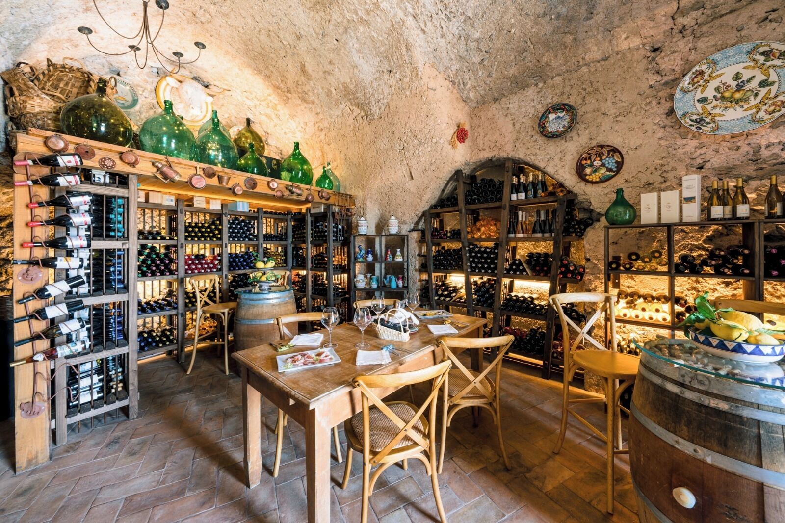 A wine cellar with wooden racks and barrels, a dining table with chairs, and a rustic stone ceiling.