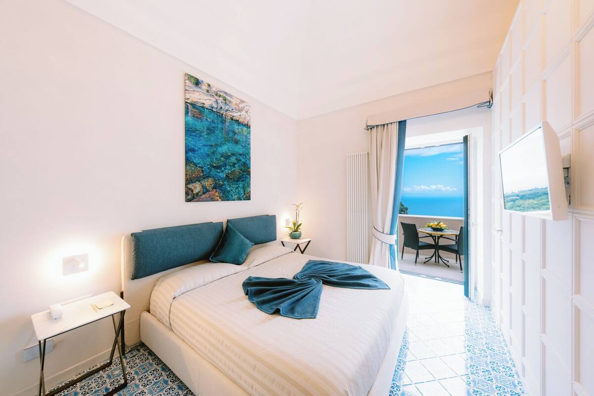 Une chambre d’hôtel avec un lit, un tableau représentant un paysage côtier, une petite table avec une plante et un balcon avec une table et des chaises.