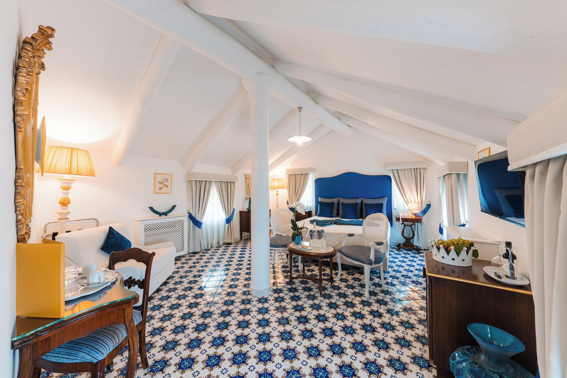 A hotel room with a high ceiling, a bed with a blue headboard, a dining area with a table and chairs, and a patterned floor.