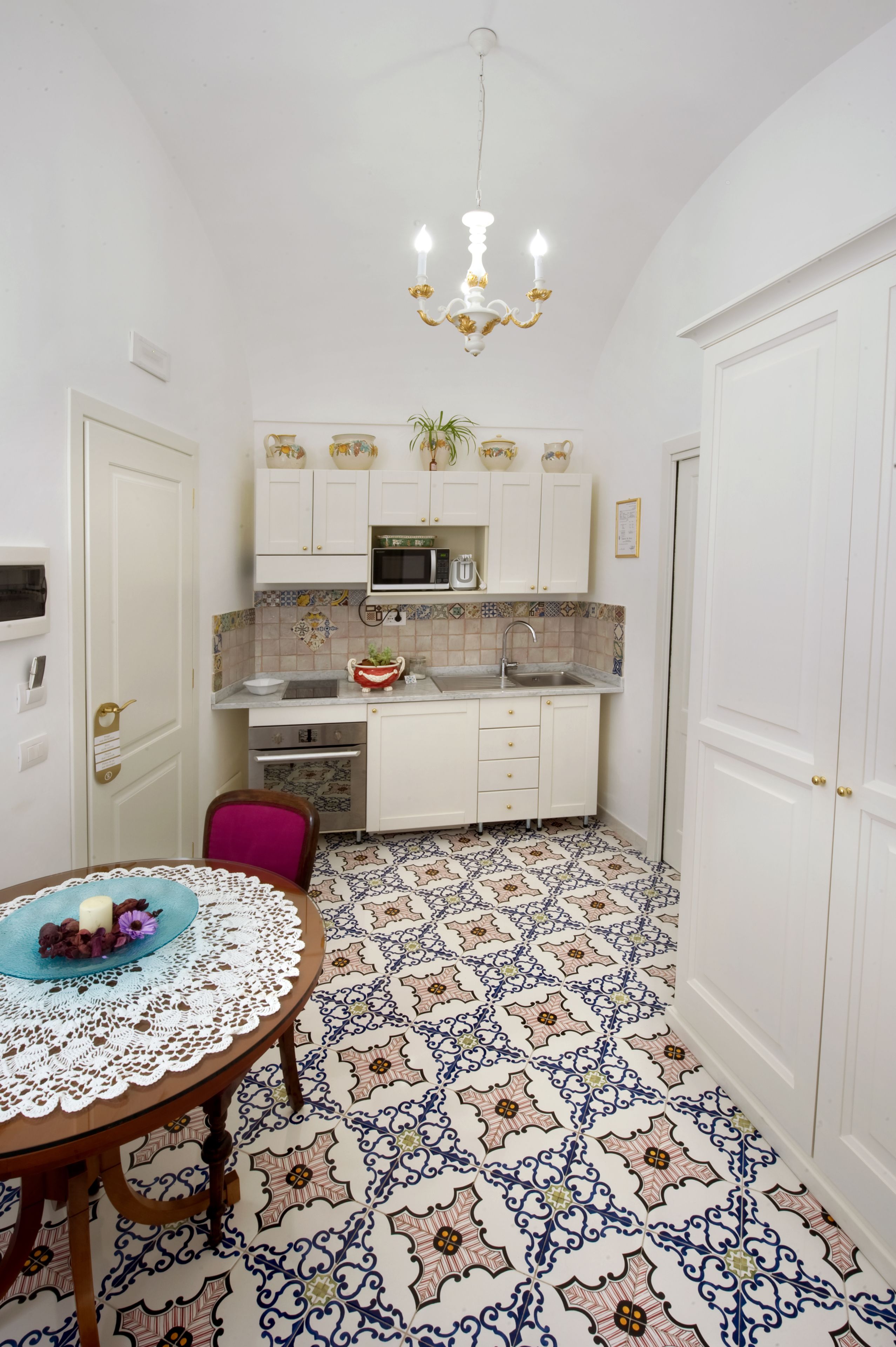 Une petite cuisine avec des meubles blancs, un four à micro-ondes et un évier. On y trouve une table recouverte d’une nappe en dentelle et une chaise. Le sol est décoré de carreaux formant un motif.