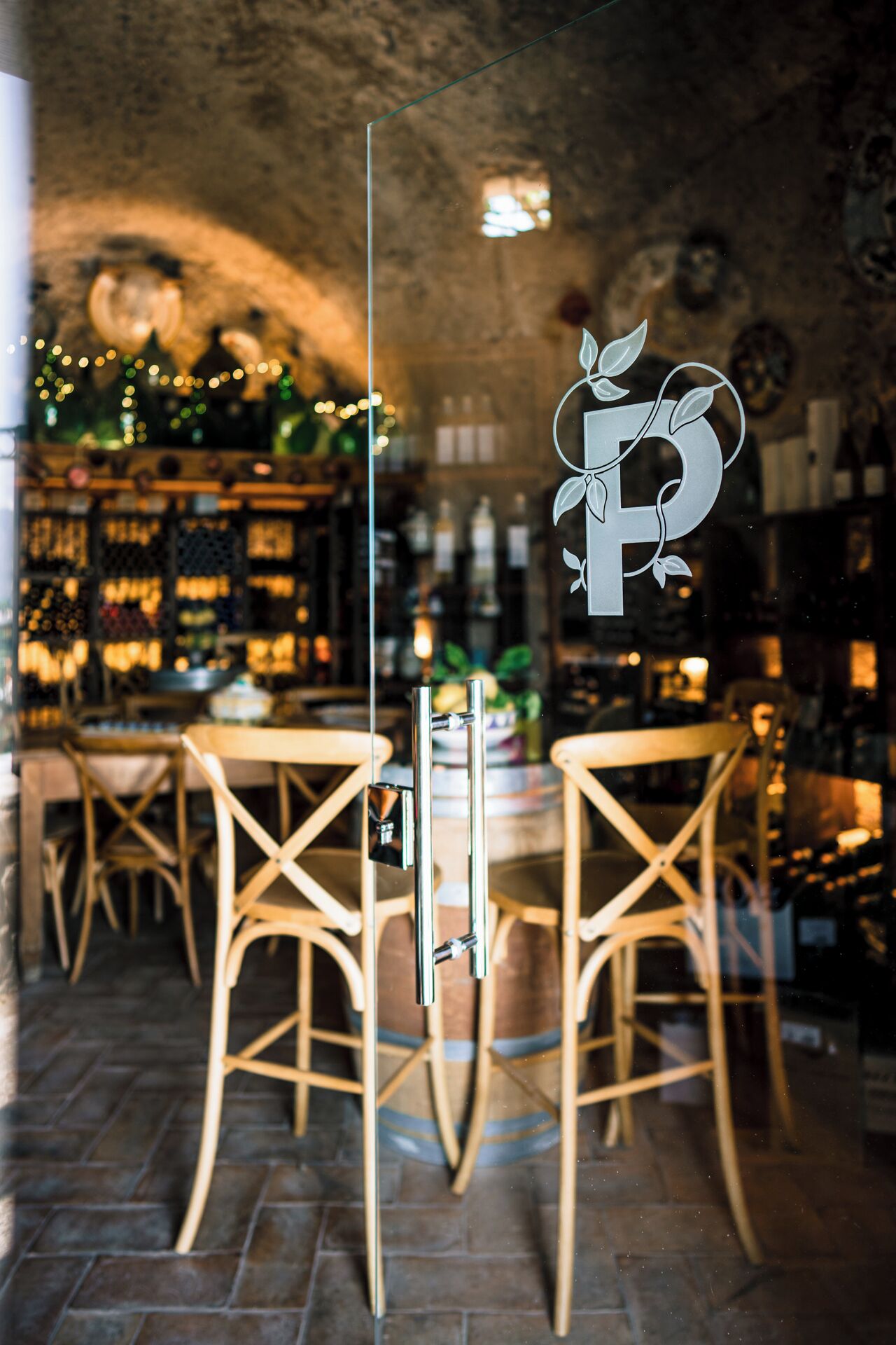 A rustic interior with a stone wall, wooden chairs, a bar area with bottles, and a decorative letter 'P' on the glass.