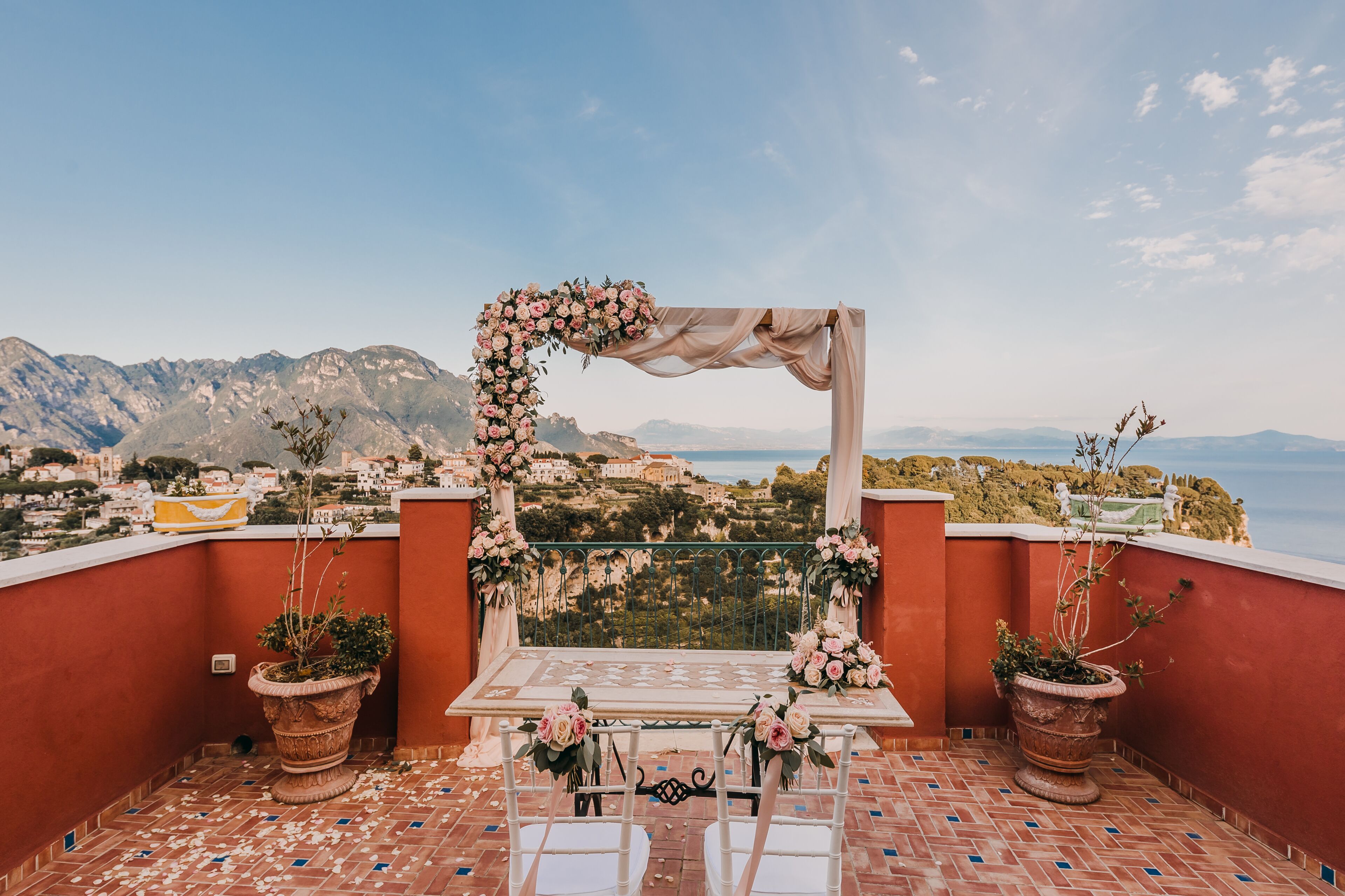 Terraço com mesa e cadeiras, decorado com flores e um arco coberto por tecido, com vista para uma paisagem montanhosa deslumbrante.