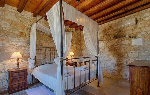 A four-poster bed with white drapes, a stone wall, a wooden ceiling, and a nightstand with a lamp. - Kirithra (Neo Chorio)