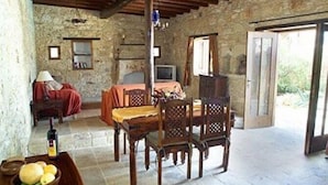 A rustic room with stone walls, a wooden table, a bed with a red blanket, and a window with a view of greenery. - Kipseli (Neo Chorio)