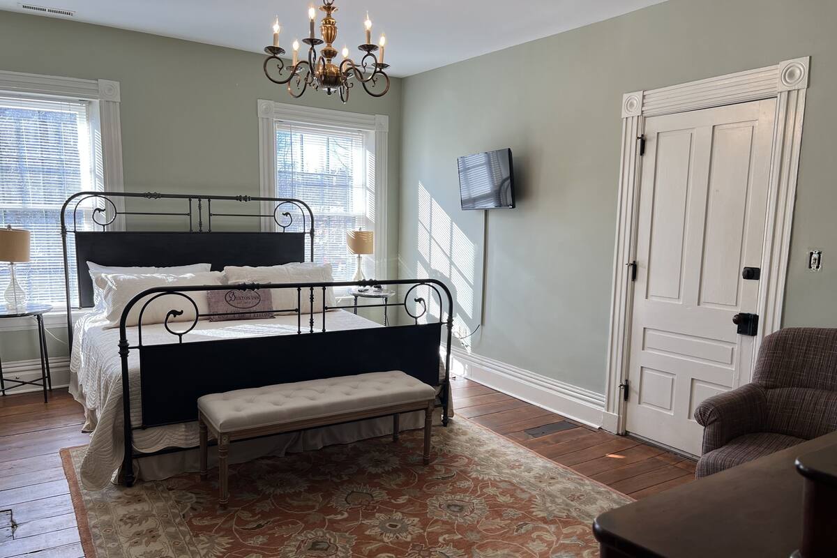 Une chambre à coucher avec un lit en métal, un banc, un lustre, un téléviseur fixé au mur et une porte.