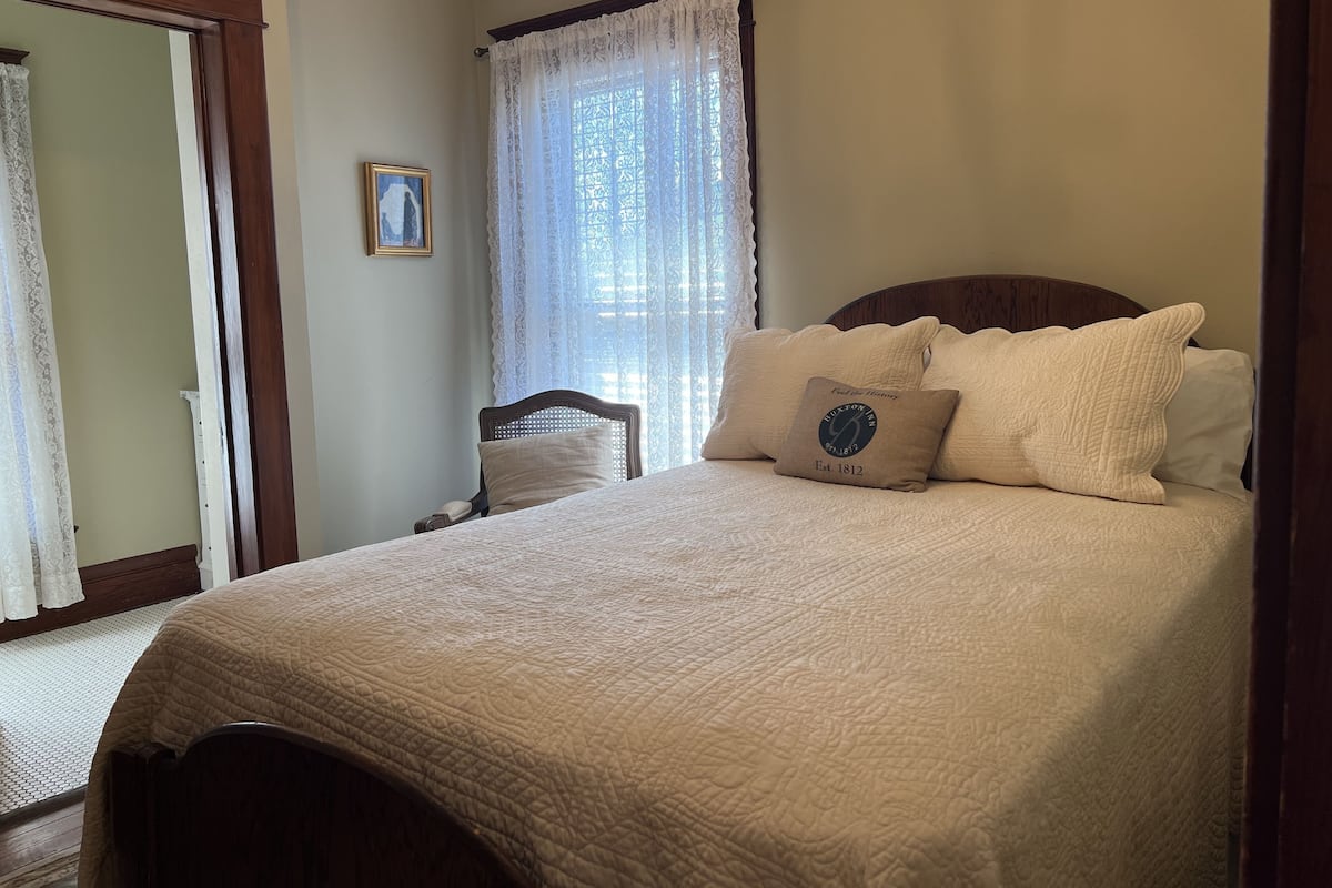 Une chambre à coucher bien rangée, avec un grand lit, une tête de lit en bois, un tableau encadré au mur et une fenêtre ornée de rideaux en dentelle.