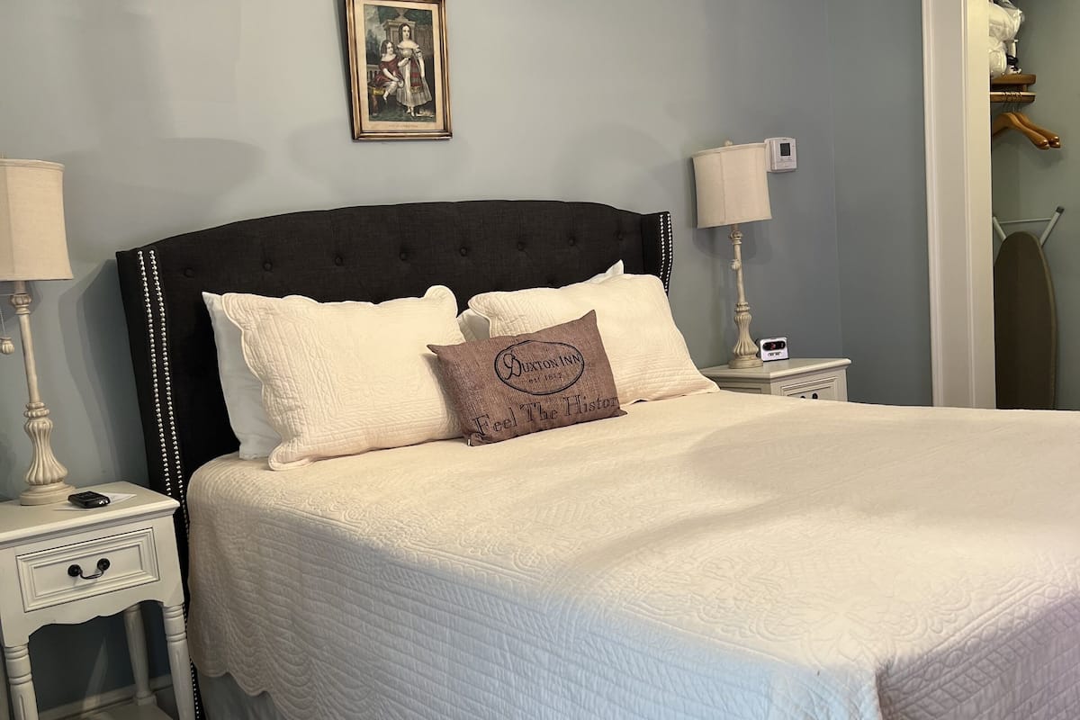 Un lit bien fait, avec une tête de lit sombre et du linge de lit blanc, une table de chevet avec une lampe et un tableau encadré au mur.