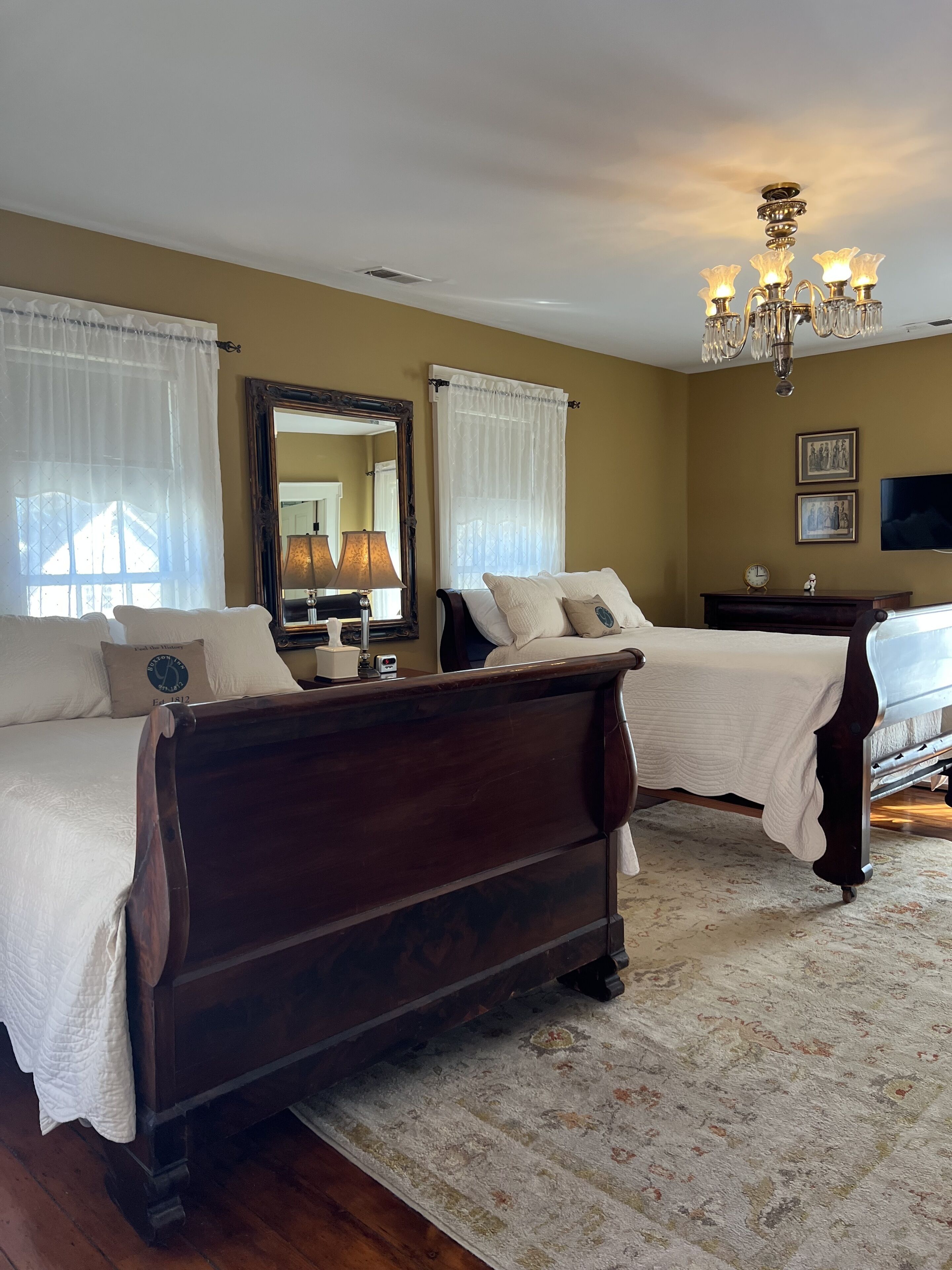 A hotel room with two wooden beds, a large mirror, a chandelier, and a television on the wall.